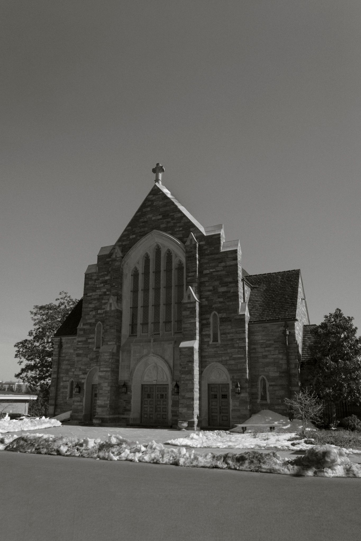 Black and white photo of a historic stone church with Gothic architecture, cross on peak, arched windows, and snow on the ground.