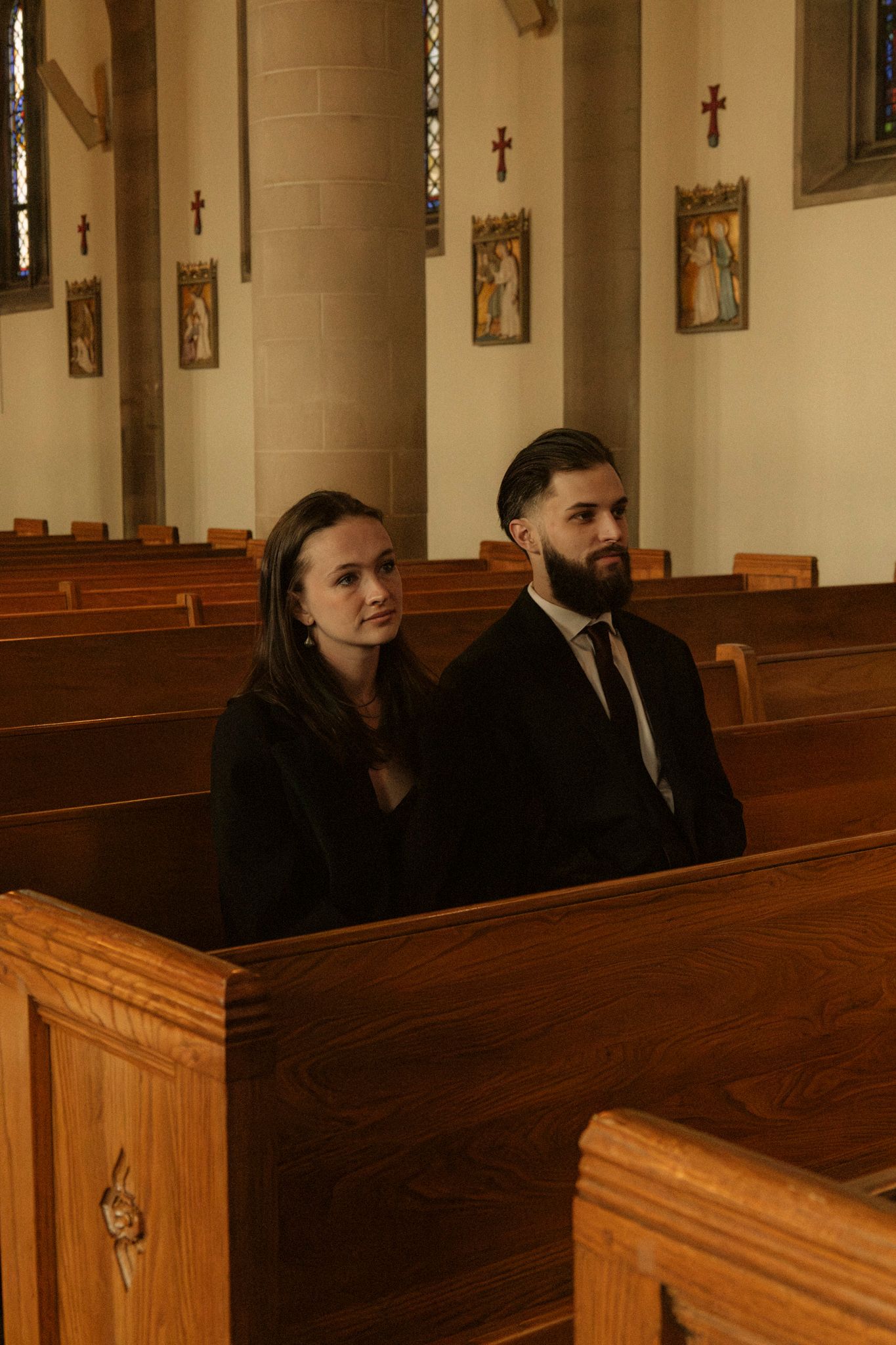 A couple dressed in black sit together in wooden church pews during a wedding ceremony.