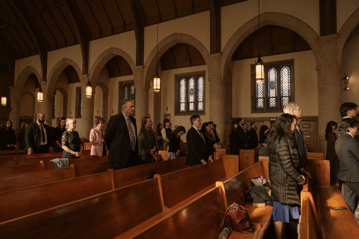 Congregation standing in wooden pews inside a historic stone church with arched ceilings and stained glass windows.