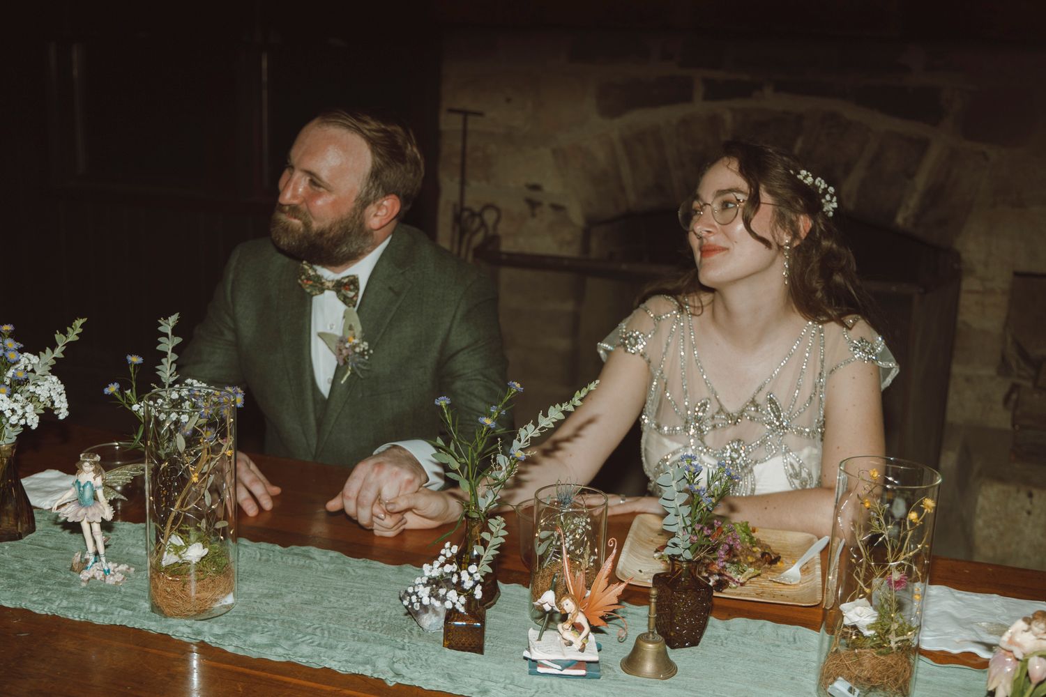 Wedding guests seated at a rustic wooden table during an intimate reception dinner with soft lighting and floral decor.