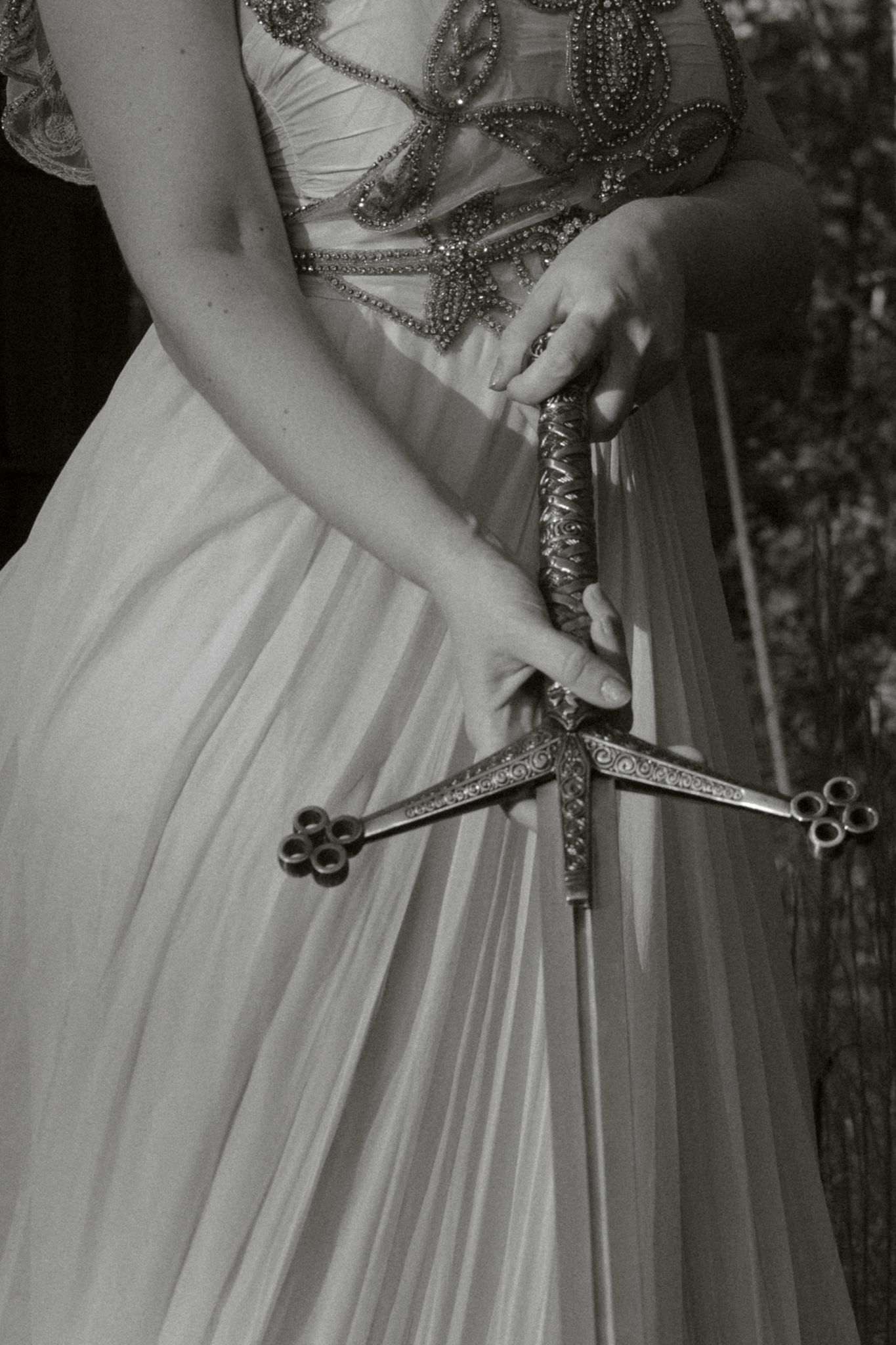 A decorative sword is held against a flowing white gown with beaded details in a black and white photograph.