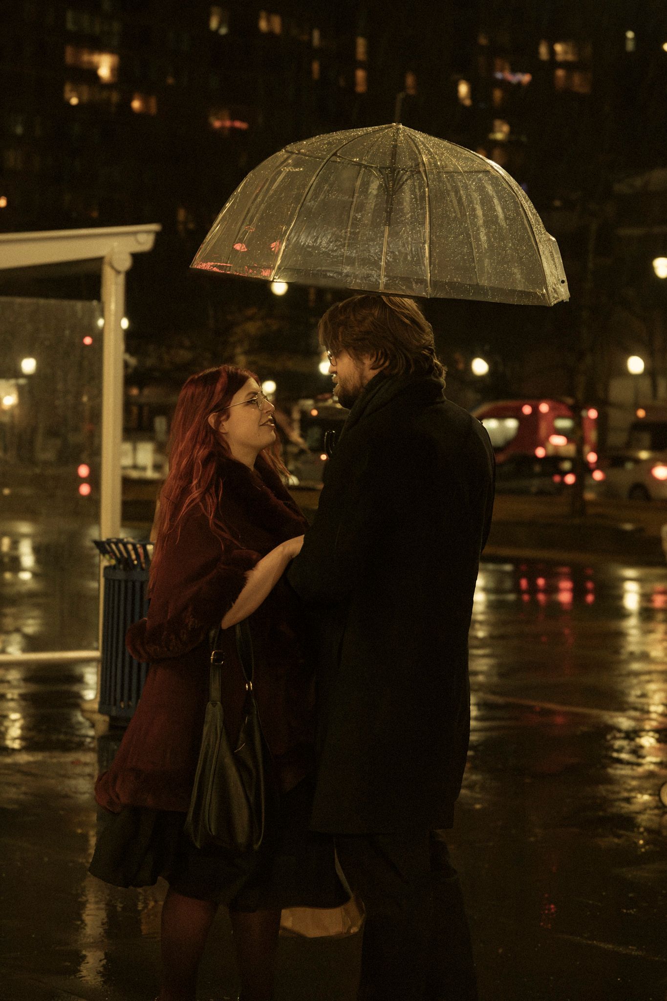 A couple shares an intimate moment under a clear umbrella at night in the rain on a city street.
