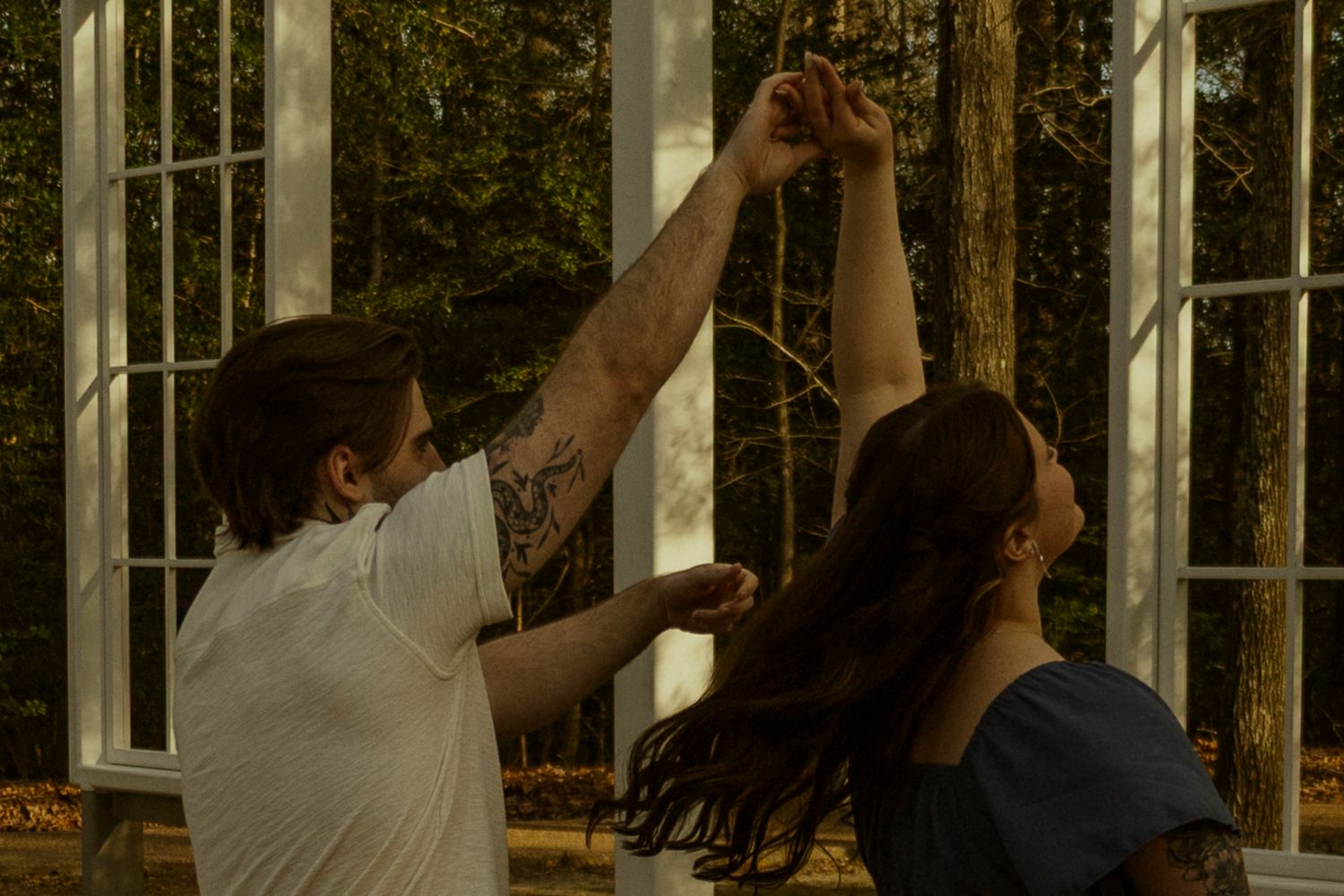 A tattooed man spins a woman with long dark hair near large white-framed windows with trees in background.