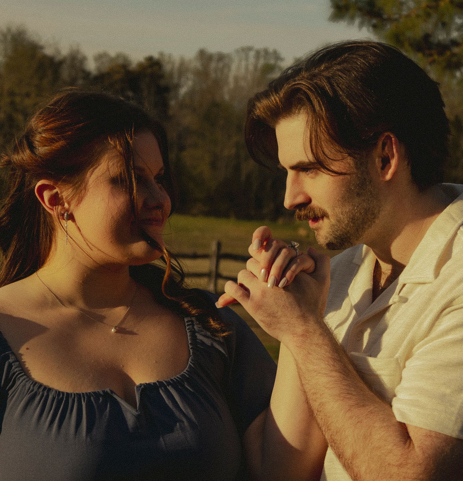 Couple holding hands intimately at golden hour near a wooden fence in a rural setting.
