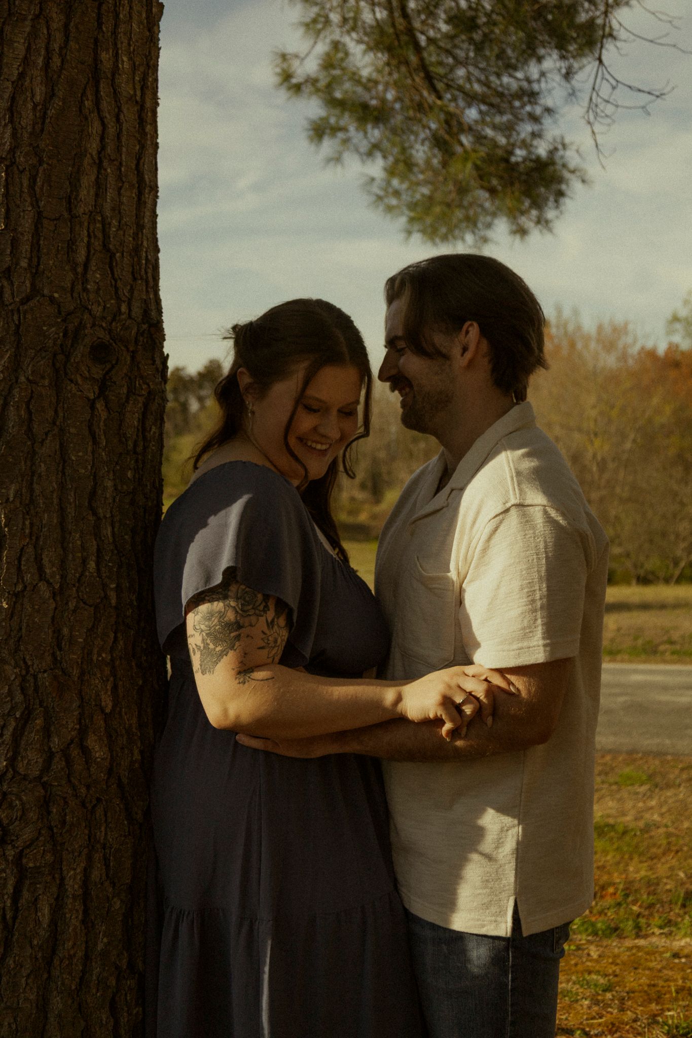 Couple holding hands and smiling near a tree in golden hour sunlight outdoors.