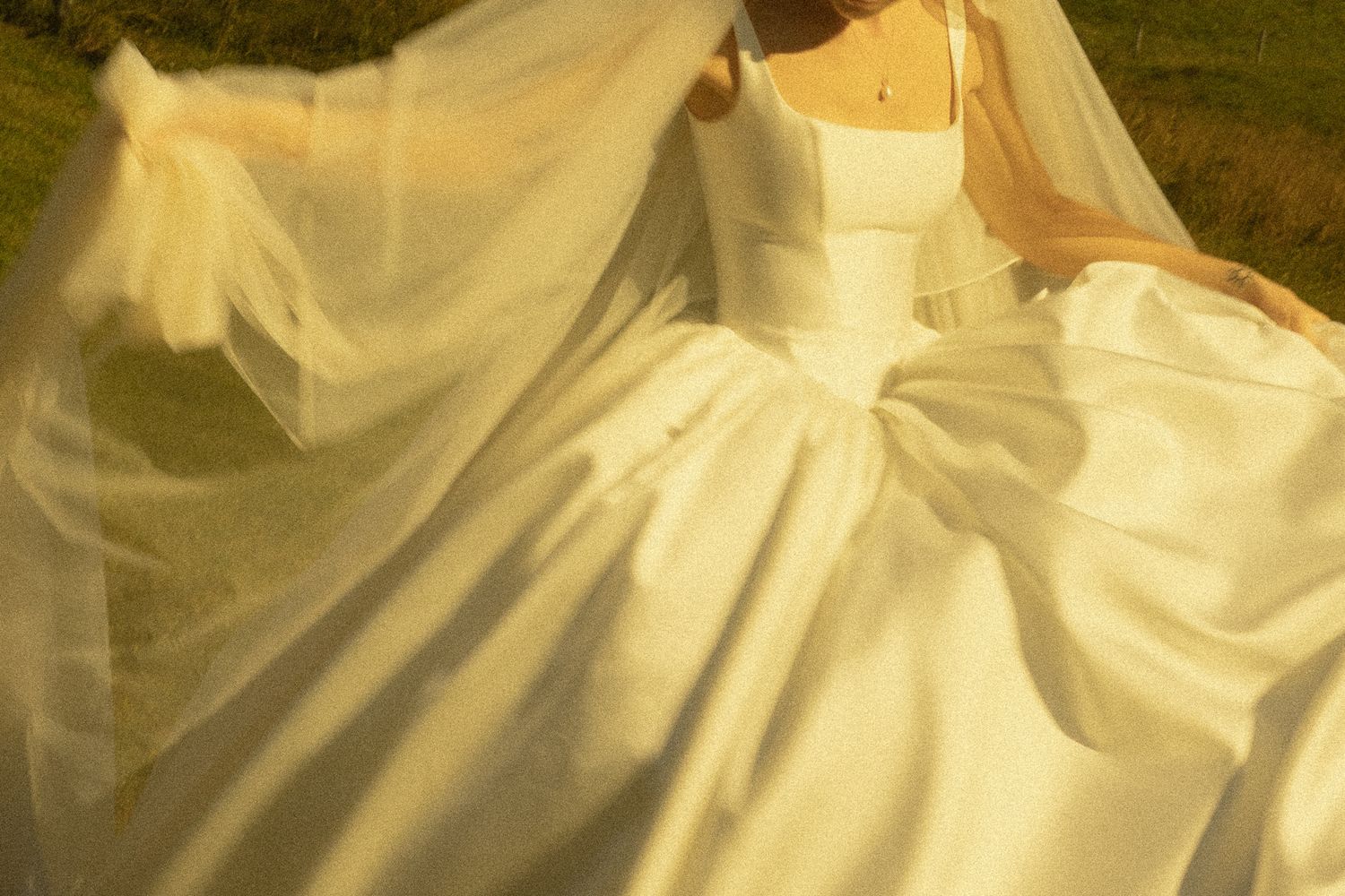 Bride in a white satin ball gown with a bow detail and flowing sheer veil, bathed in warm golden sunlight outdoors.