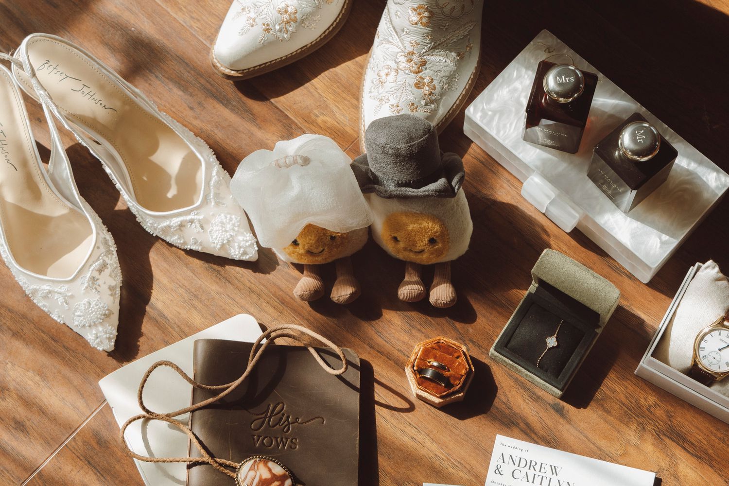 Wedding accessories including white lace shoes, rings, and decorative items arranged on wooden surface in warm lighting.