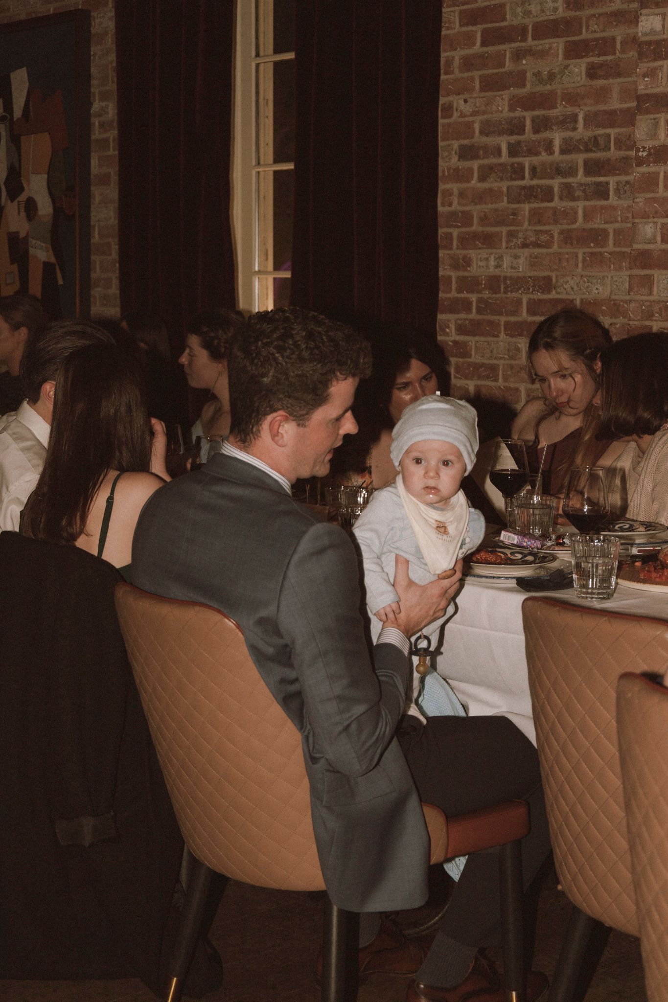 A man in a suit holds a baby dressed in white at a wedding reception dinner table.