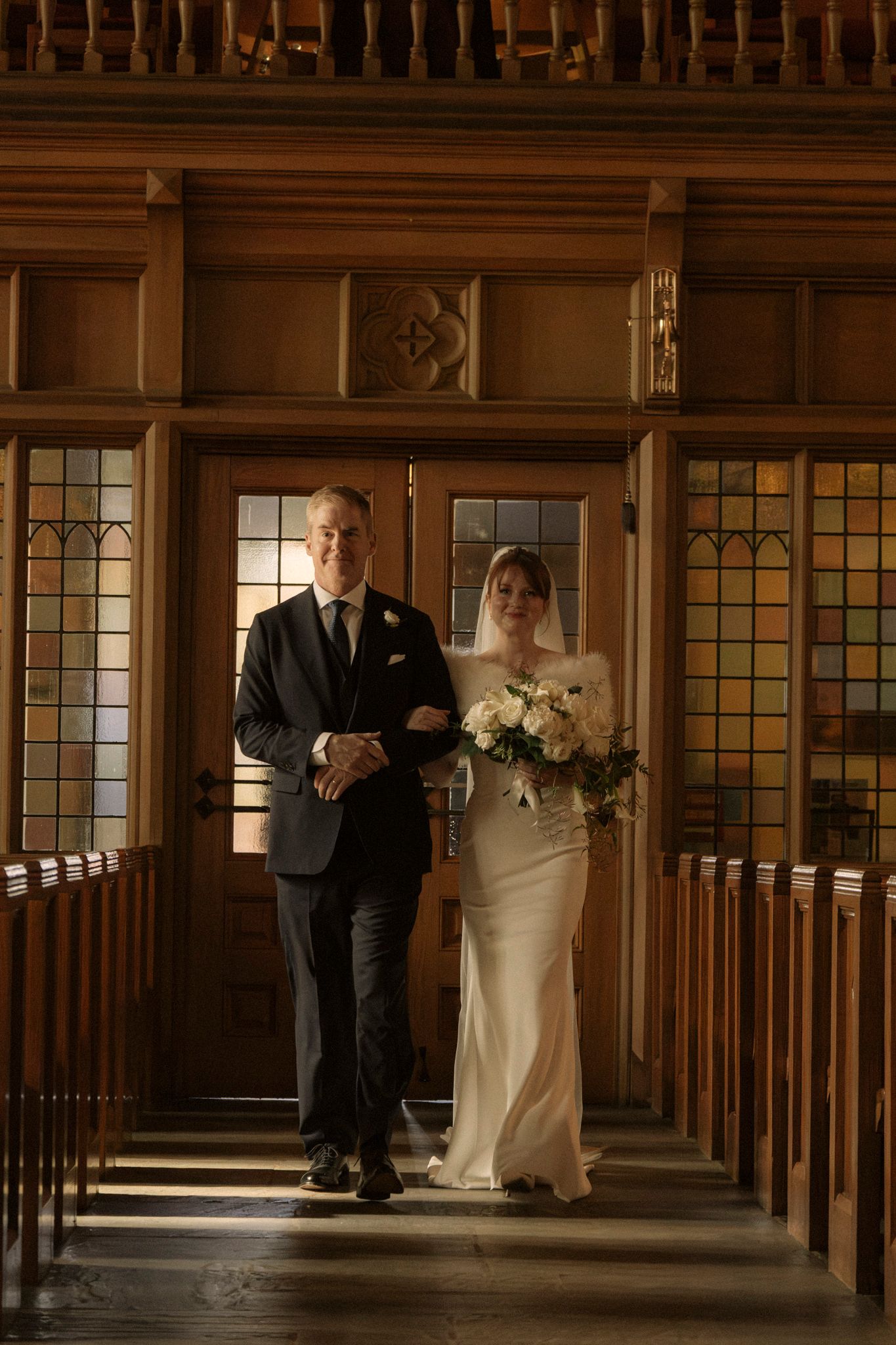 Father escorts bride in elegant white gown down church aisle past wooden pews during wedding.