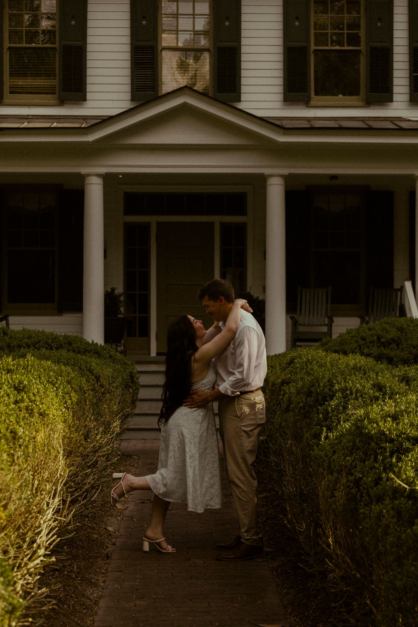 Couple sharing a kiss in front of elegant white colonial home with columns.
