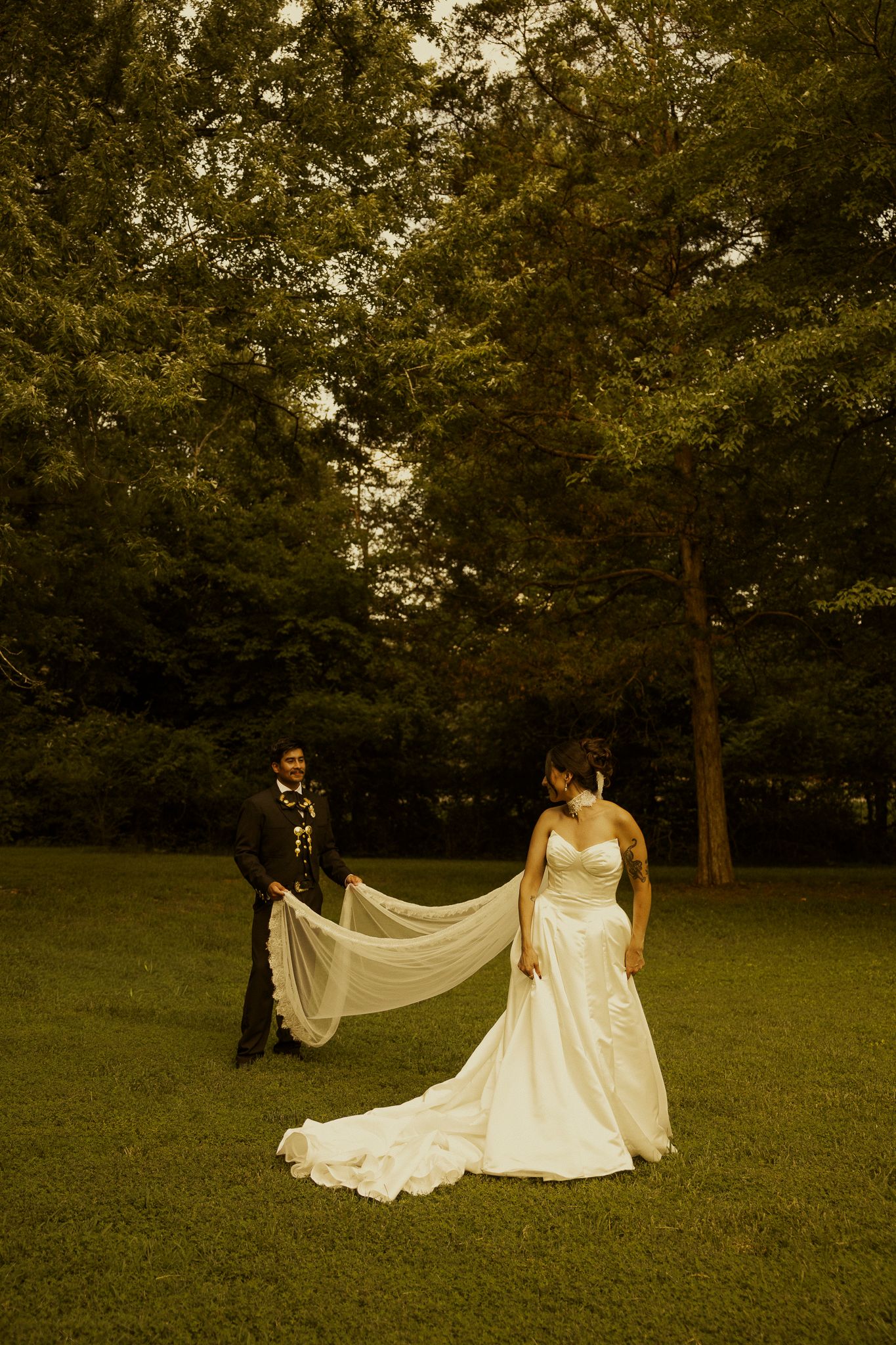 Bride in white gown with groom holding veil train in lush green park during outdoor wedding portrait.