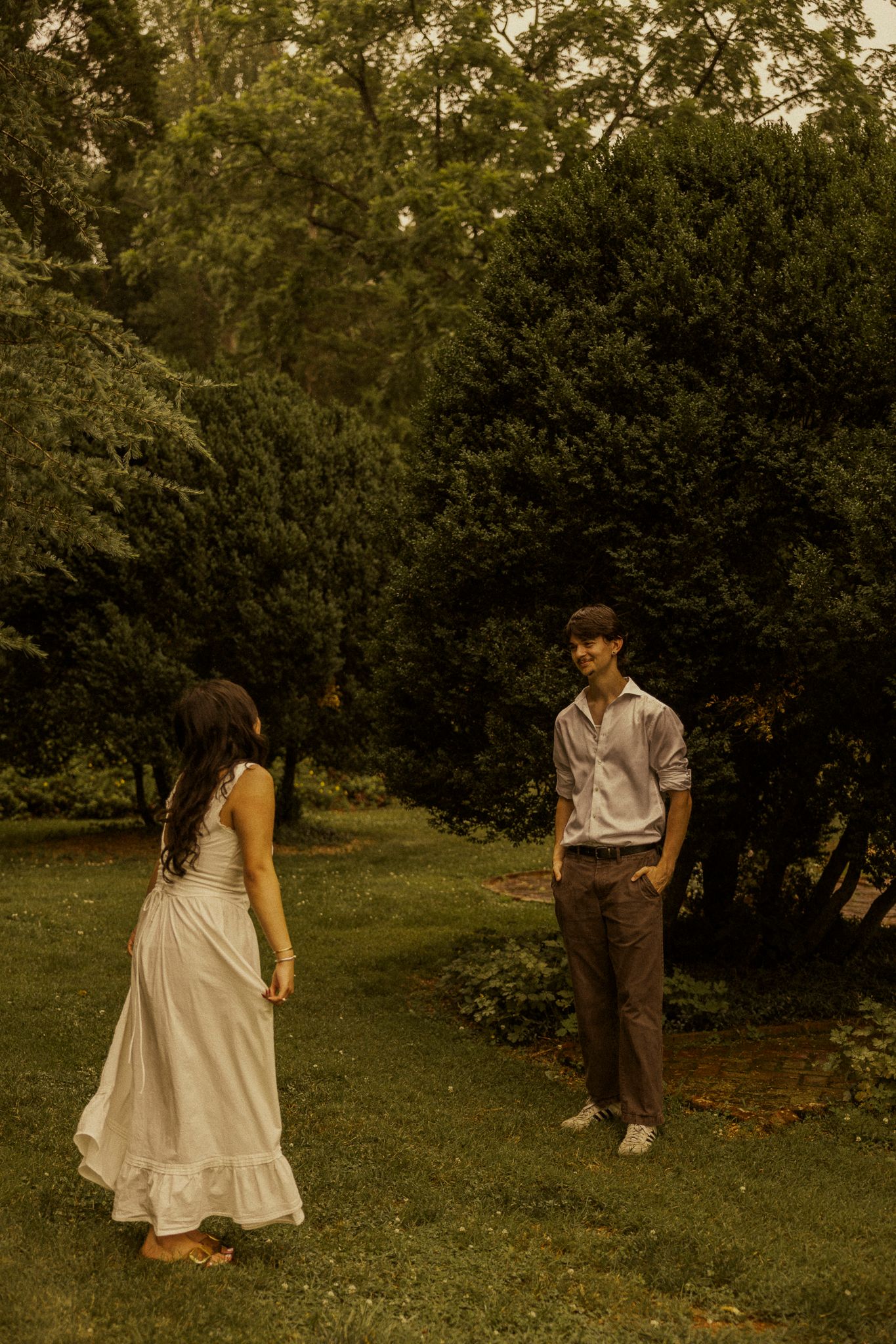 A young couple stands apart on lush green grass surrounded by tall evergreen trees in a romantic outdoor setting.