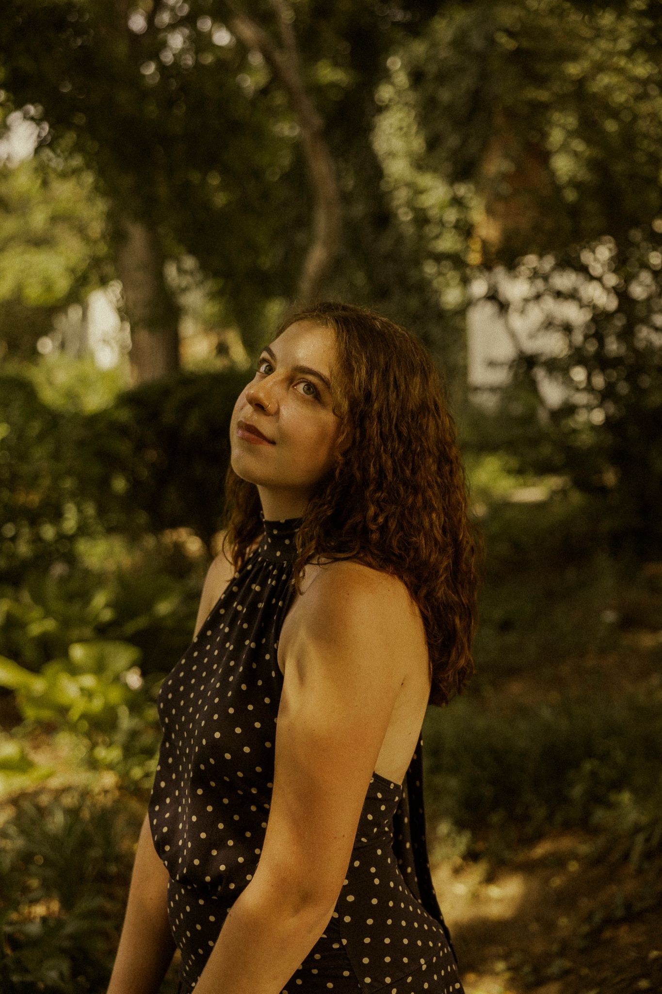 Young woman in polka dot dress gazing upward in a lush green garden setting.