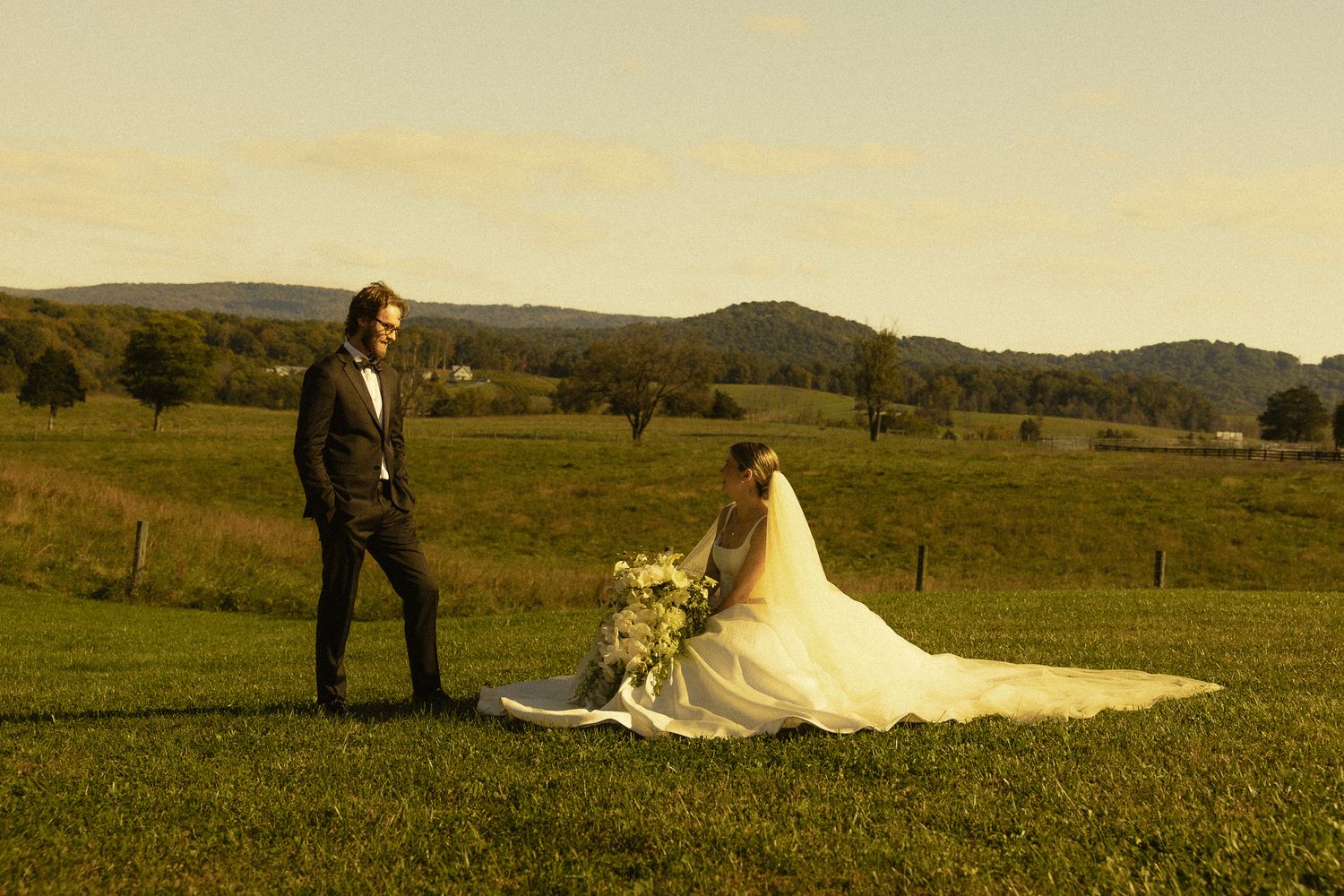 Groom in a dark suit stands beside a seated bride in a flowing white gown holding a bouquet in a grassy field with rolling hills at sunset.