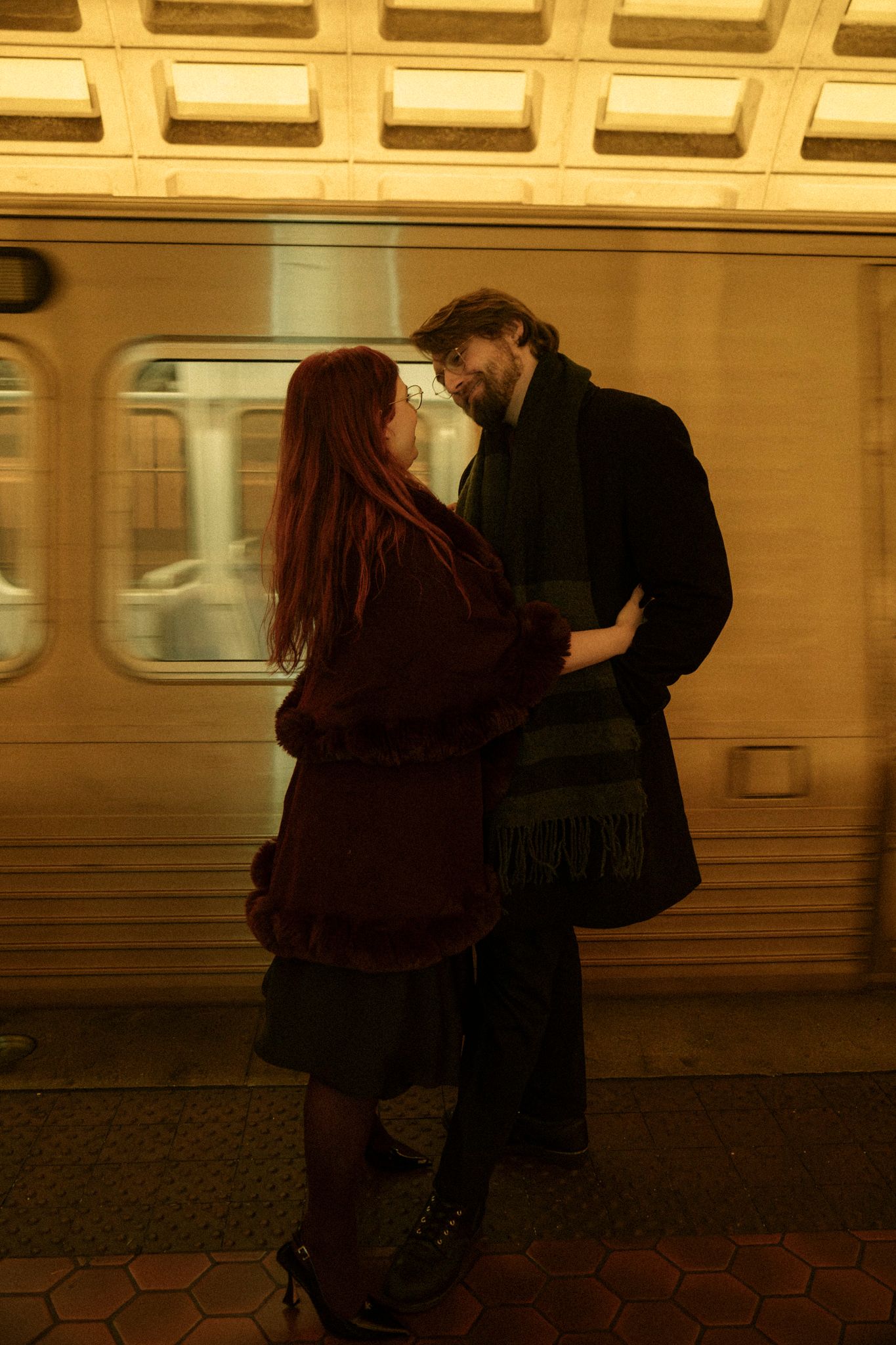 A couple embraces romantically on a subway platform with a train in the background creating a moody atmosphere.