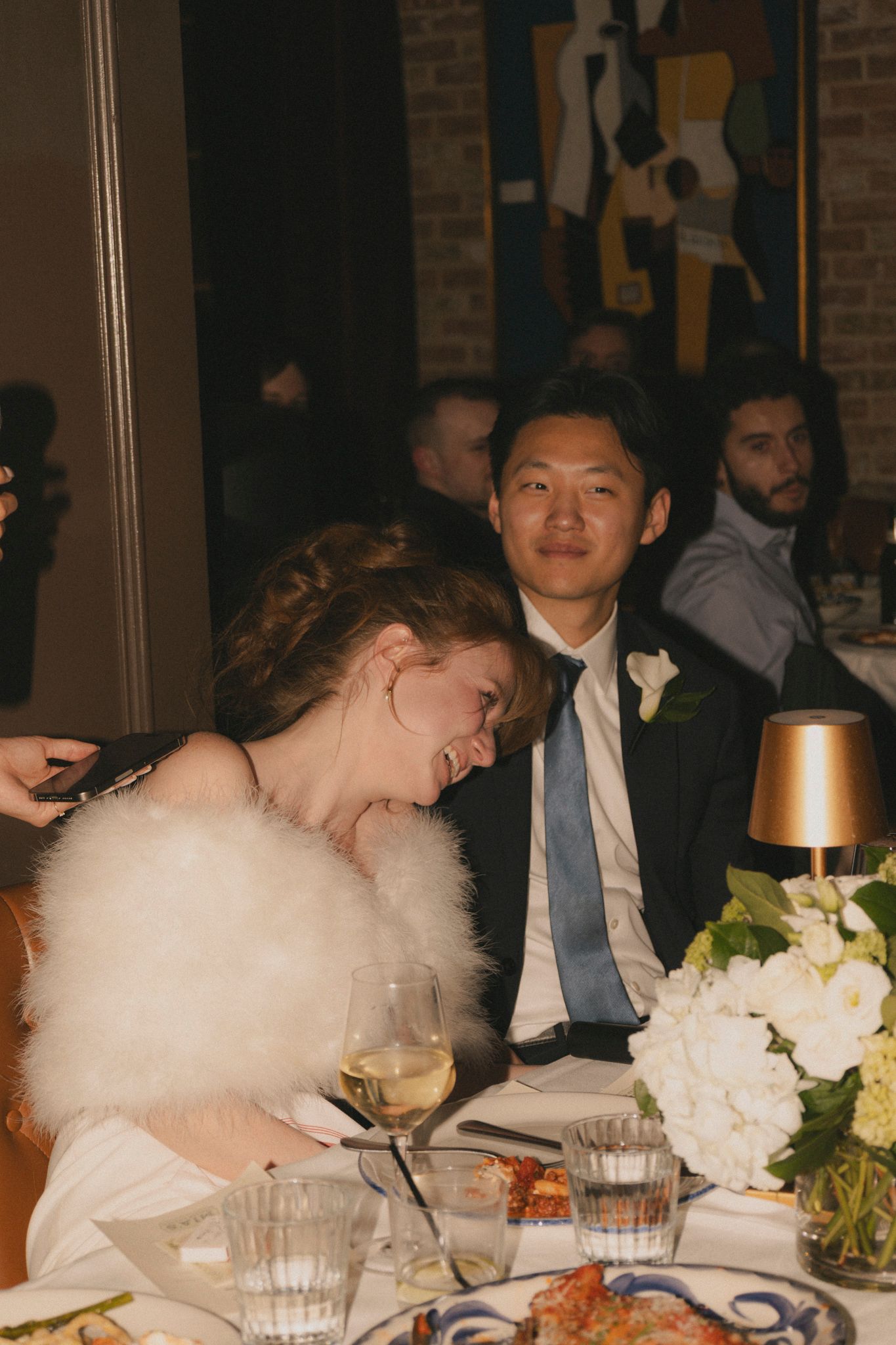 Bride in white dress with feather wrap laughing joyfully beside groom in suit at wedding reception table.