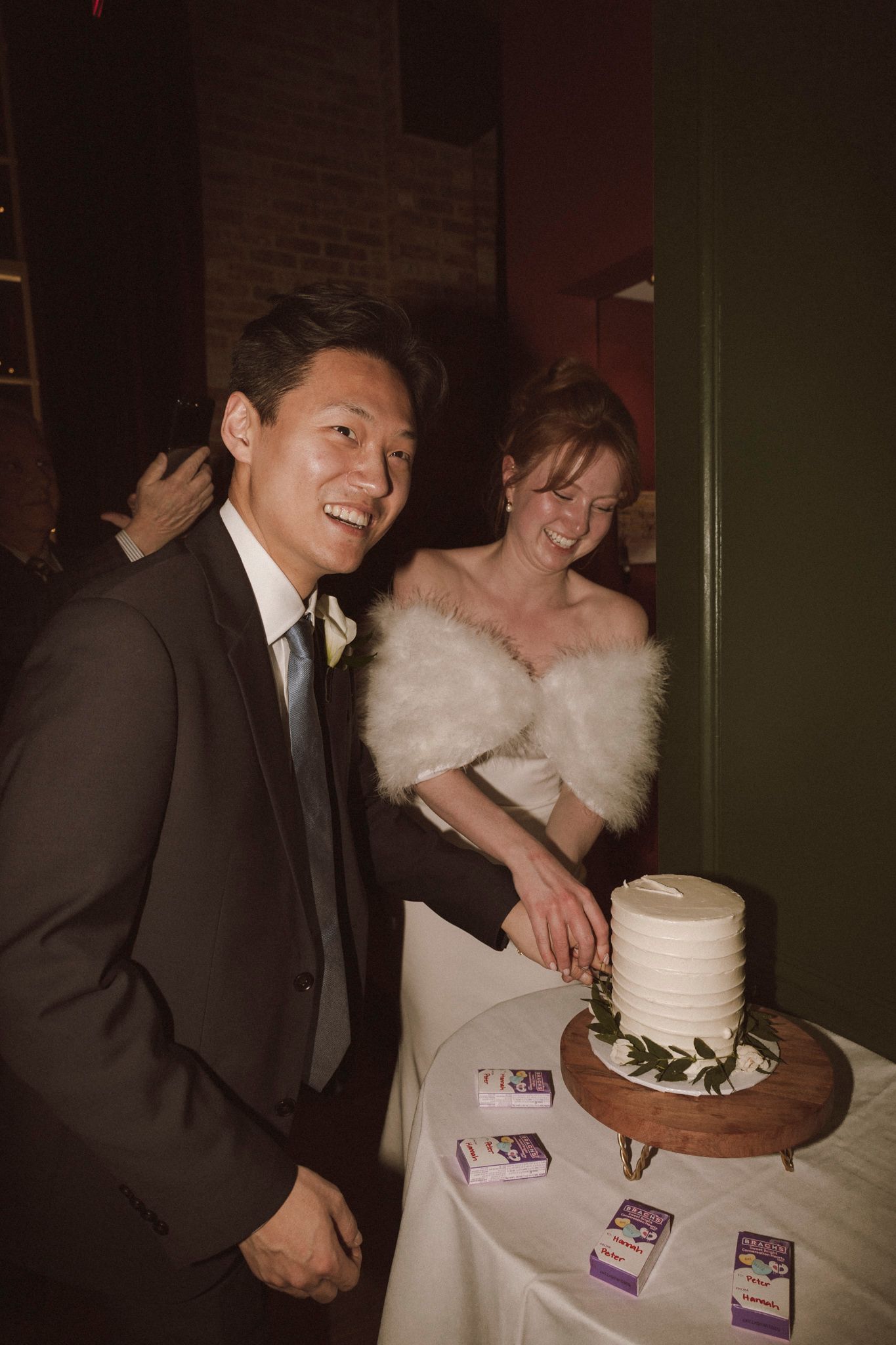 Smiling bride and groom cut their white two-tier wedding cake decorated with greenery at their reception.