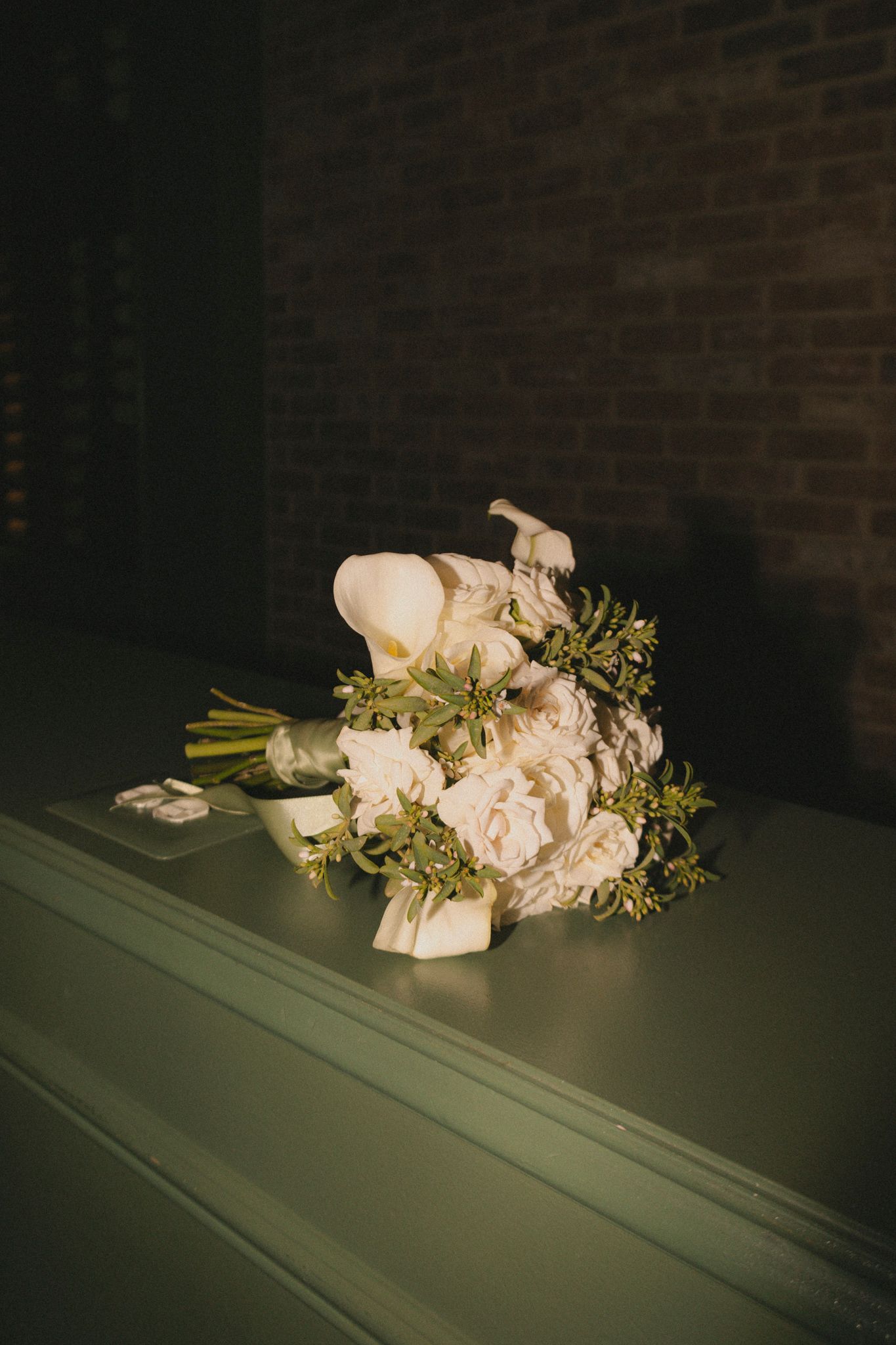 Elegant white wedding bouquet with roses and calla lilies resting on a green surface against a brick wall.
