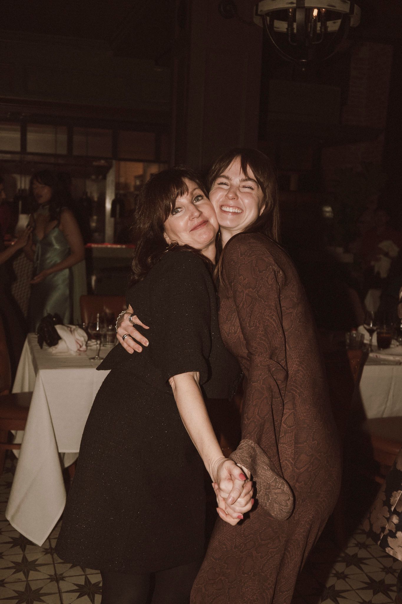 Two women smiling and embracing at a dimly lit restaurant event, holding hands and posing together.