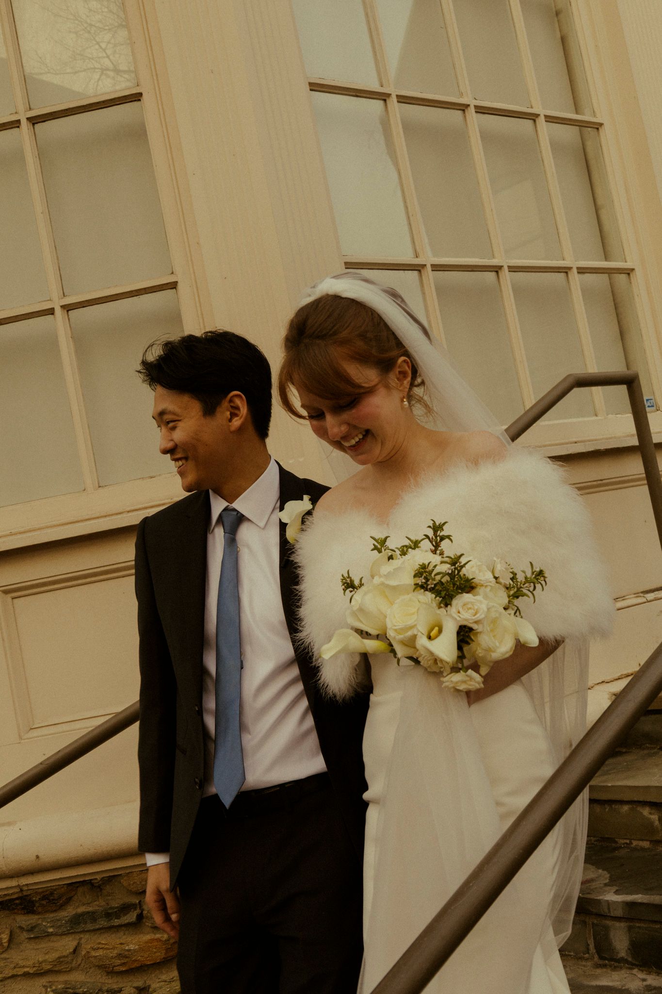 Smiling bride and groom descending stairs, bride wearing fur stole and holding white rose bouquet.