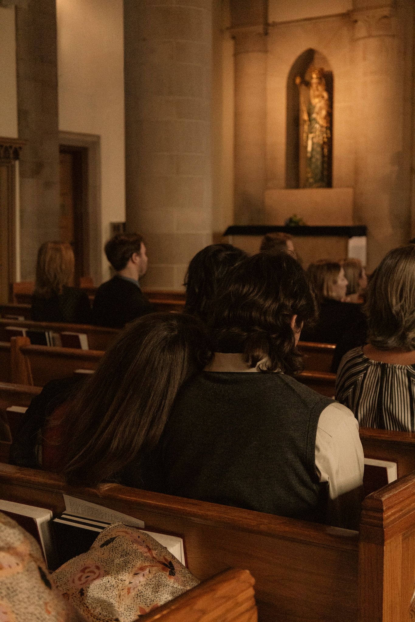 Wedding guests seated in wooden church pews facing a golden altar niche during a ceremony.