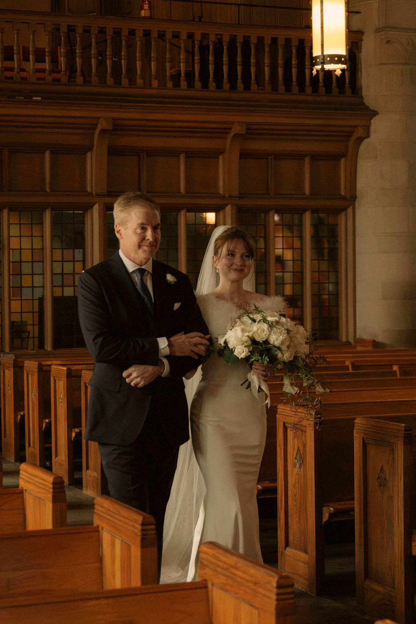 Bride in white gown walks down church aisle with escort during wedding ceremony in elegant wood-paneled chapel.