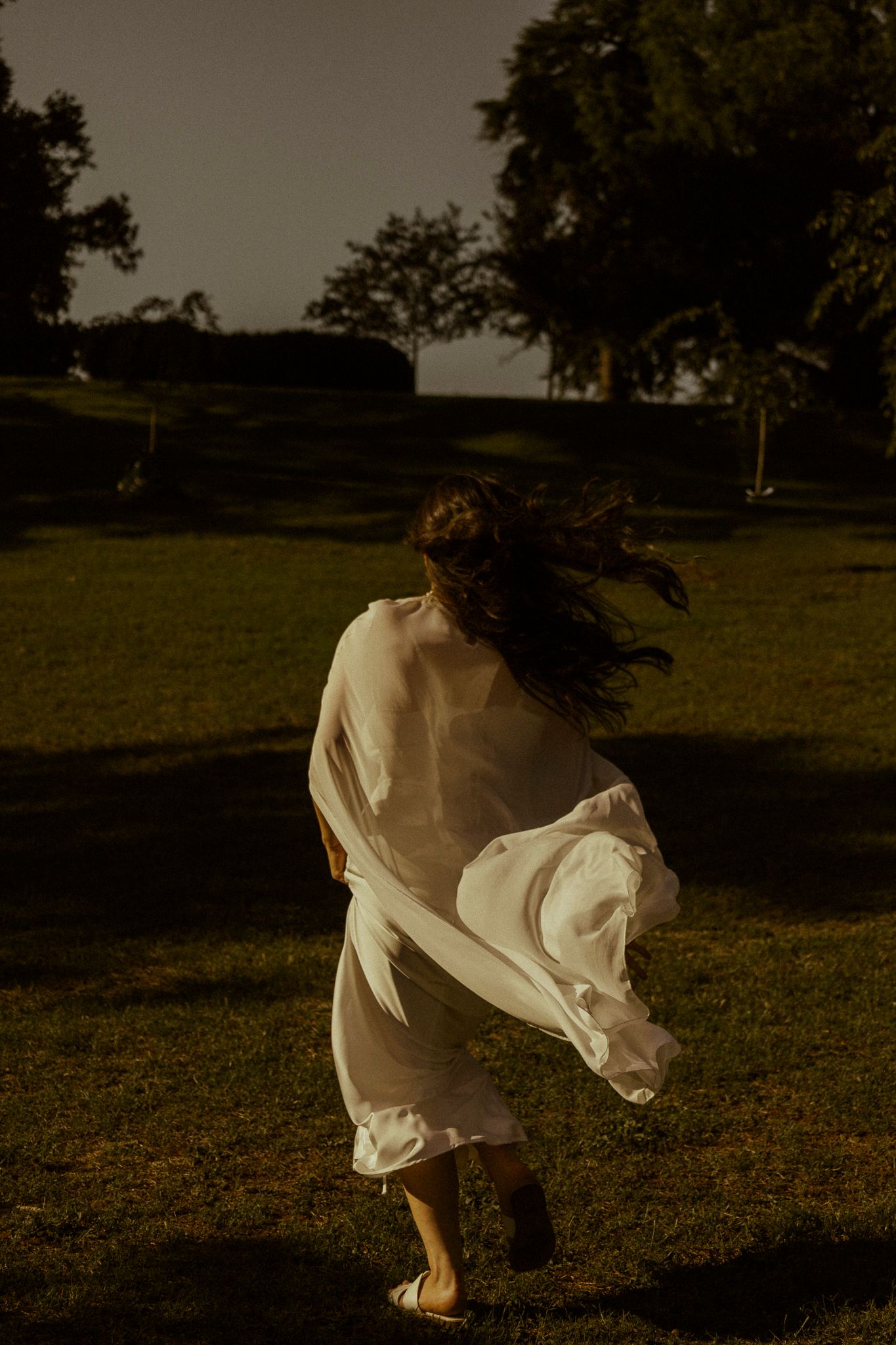 Bride in flowing white dress running across green lawn with hair blowing in golden evening light.