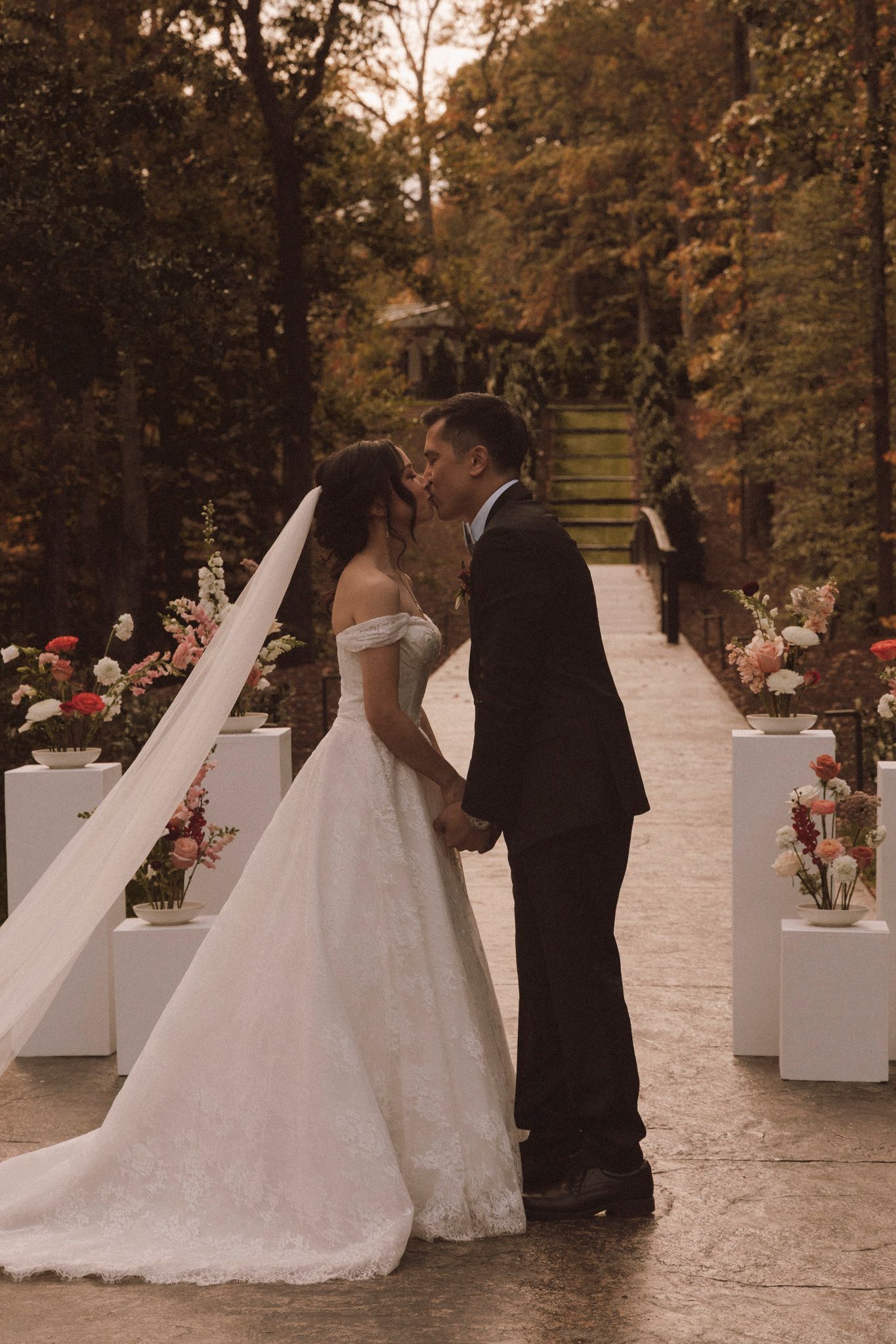 A romantic wedding scene on a garden path lined with white pedestals and pink flowers at sunset.