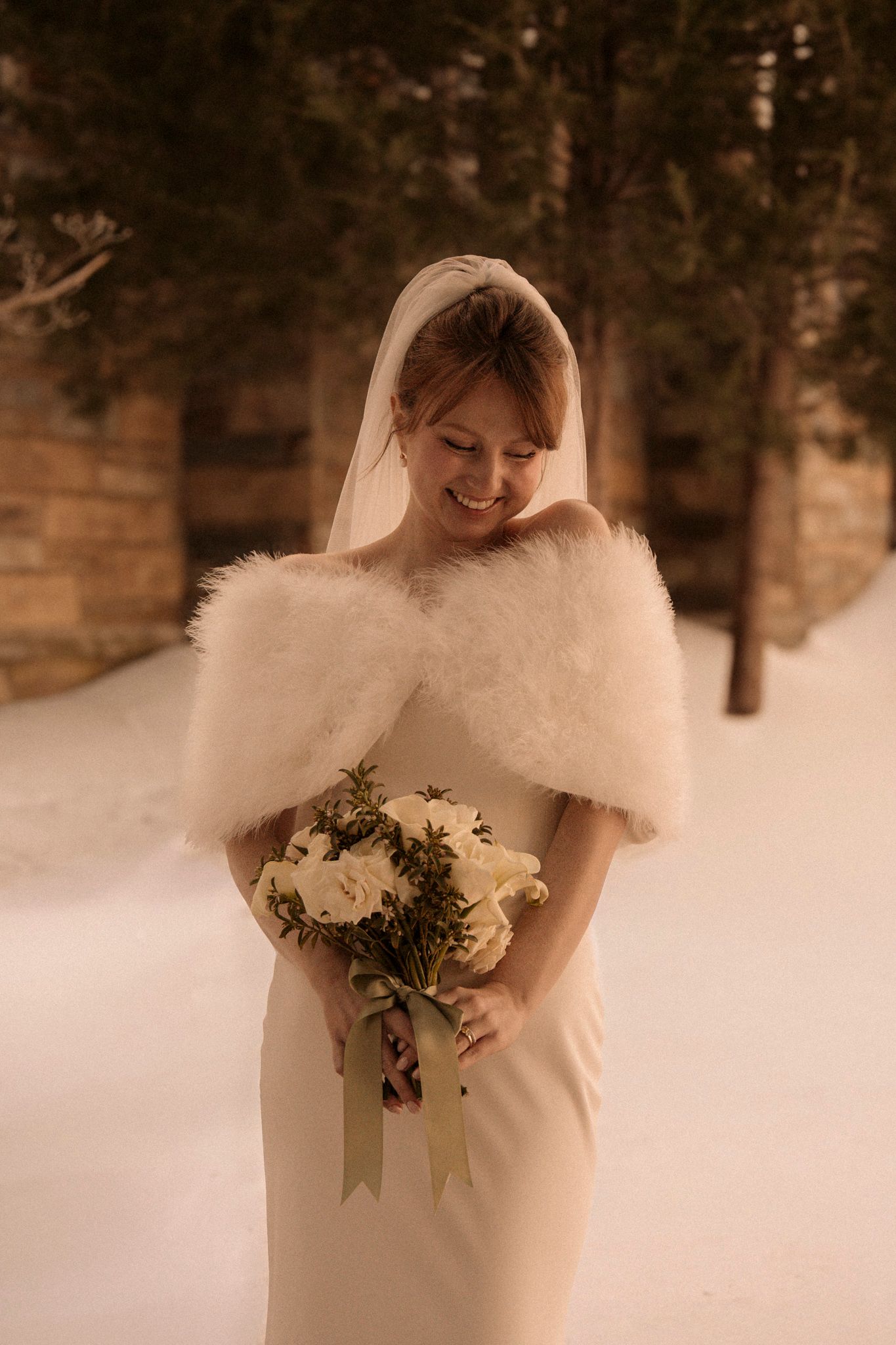 A bride in a white fur-trimmed dress holds a winter bouquet against a snowy forest backdrop.