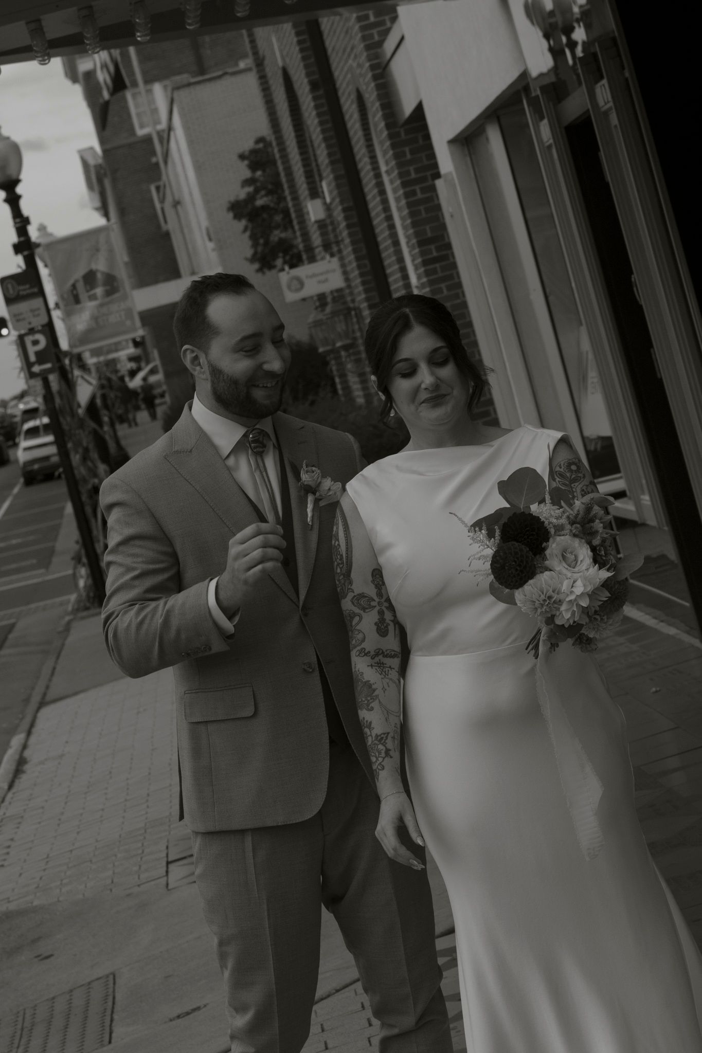 Elegant couple walking down a city sidewalk in wedding attire with the bride holding a floral bouquet.