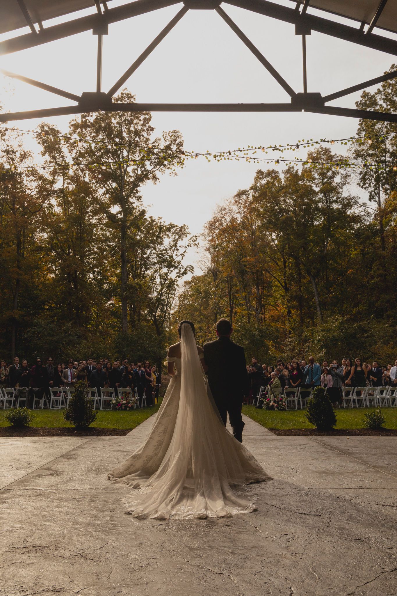A wedding couple dances under a white tent canopy during their outdoor autumn reception with guests watching in background.