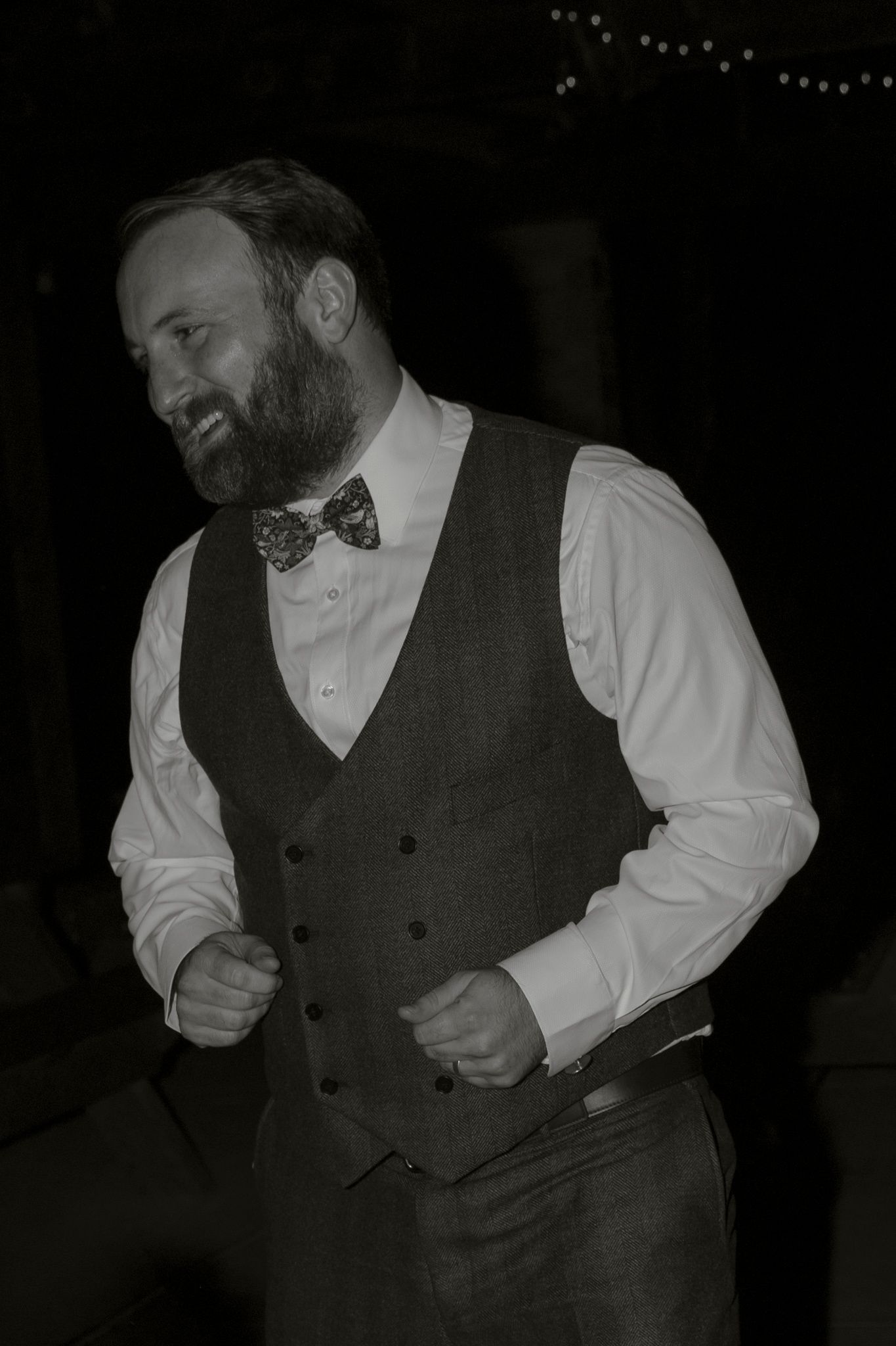 A figure in a dark vest and white shirt dances energetically in a dimly lit setting.