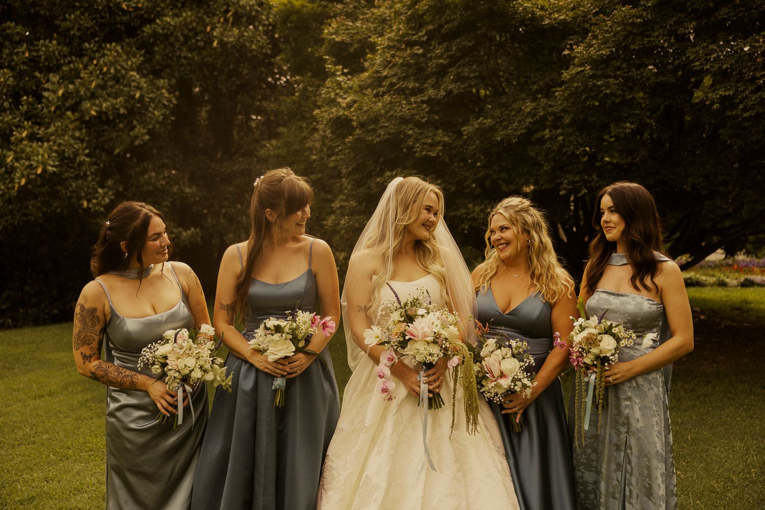 Bride in white gown with four bridesmaids in dusty blue dresses holding bouquets, smiling outdoors.