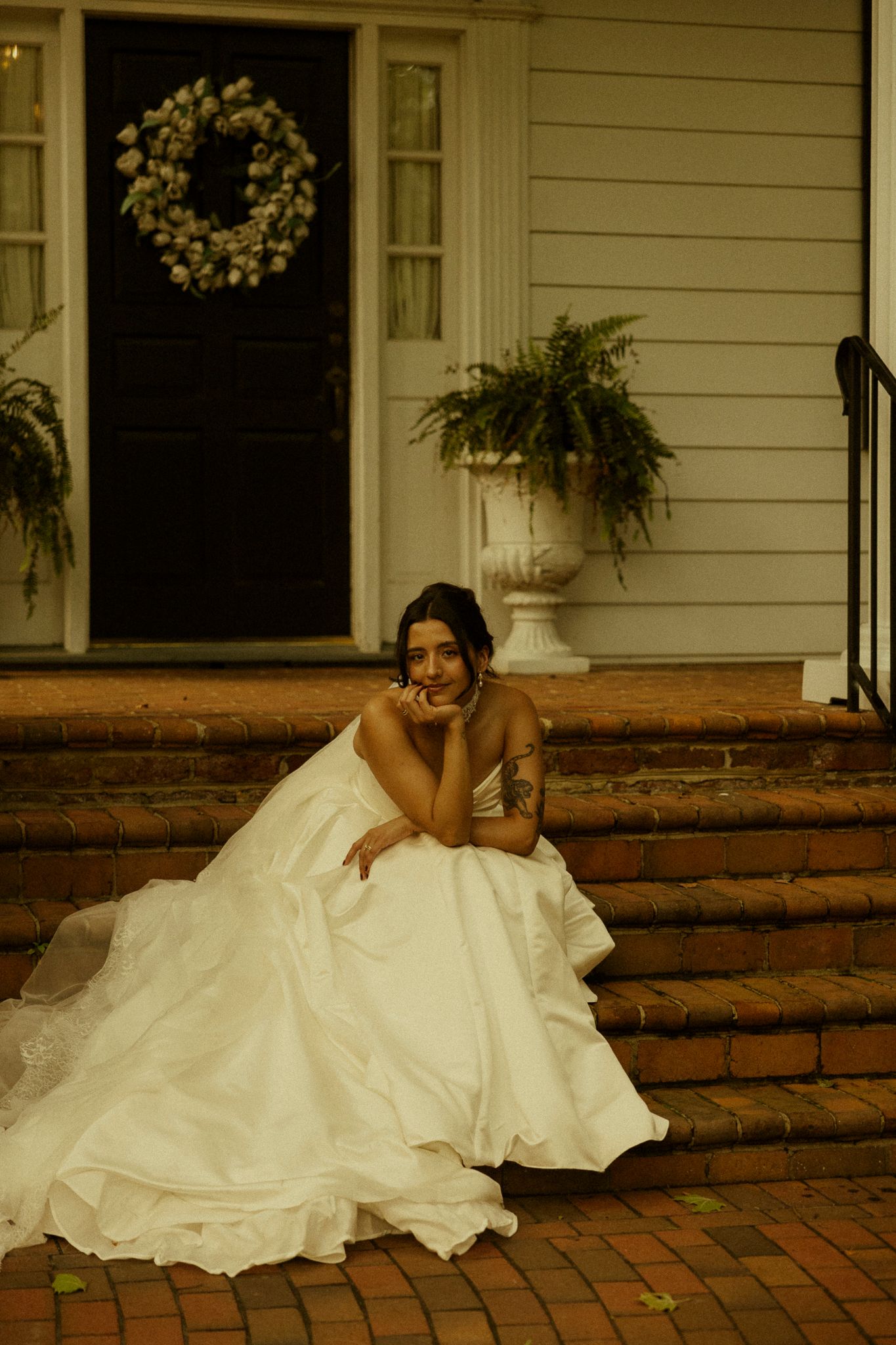 Bride in white ball gown sitting on brick steps of a classic home with wreath and ferns.