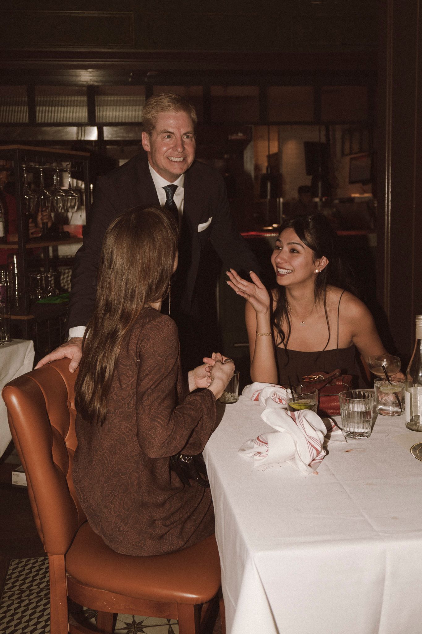 Man in black suit chats with two women seated at white-clothed table in dimly lit fine dining restaurant.