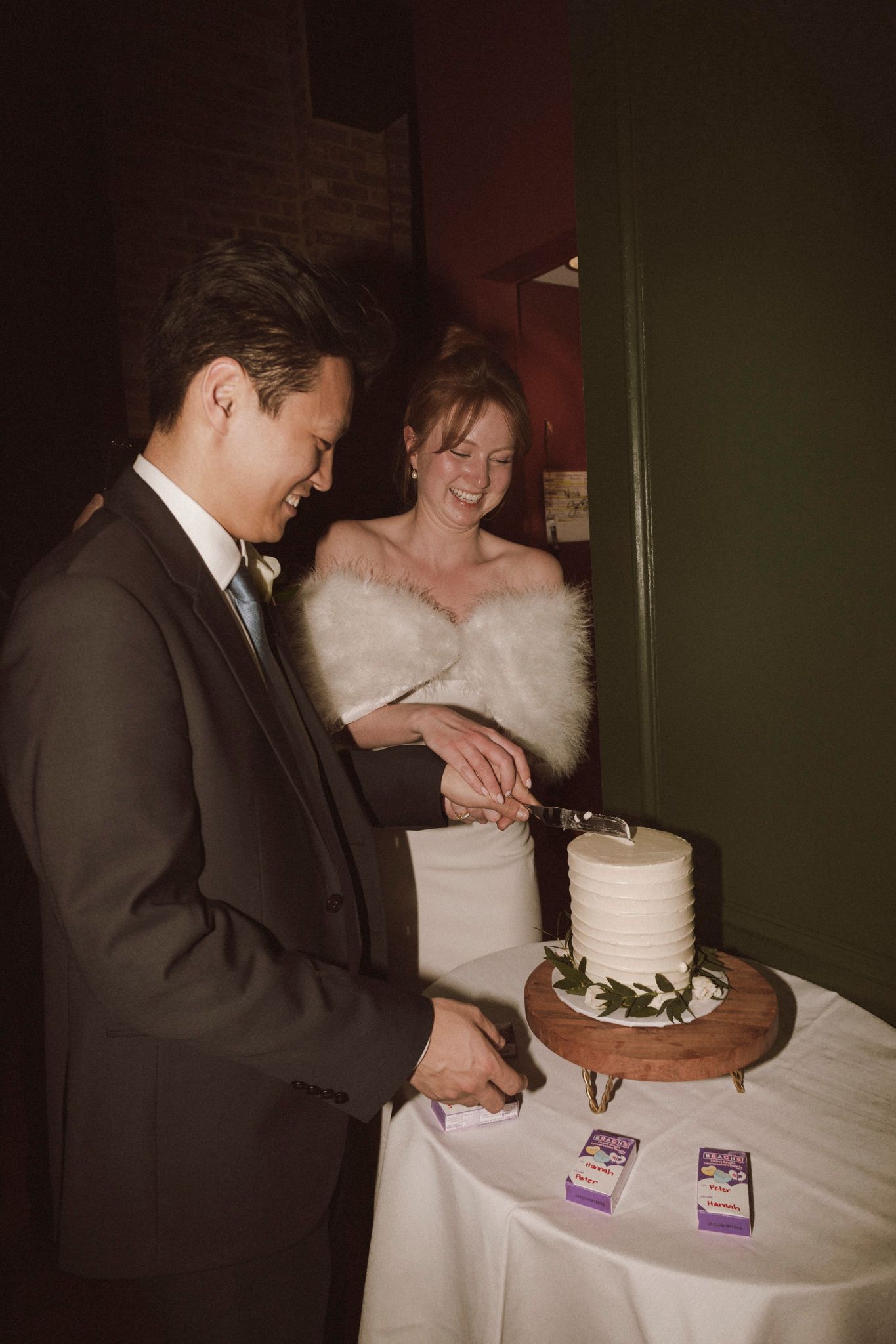 Bride and groom cutting white wedding cake together at reception, bride wearing fur stole, groom in dark suit.