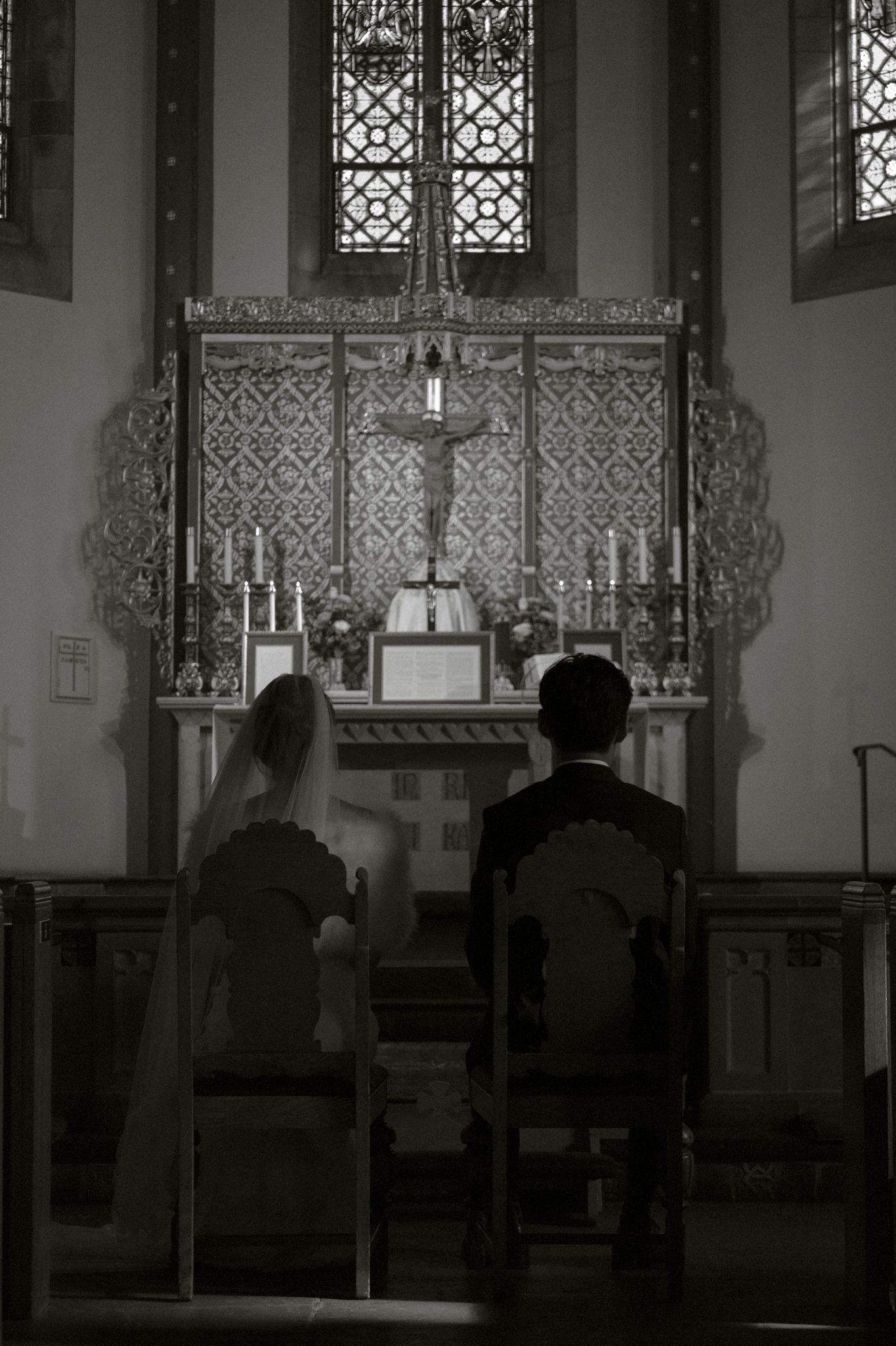 Black and white photo of bride and groom kneeling before ornate church altar with crucifix during wedding ceremony.