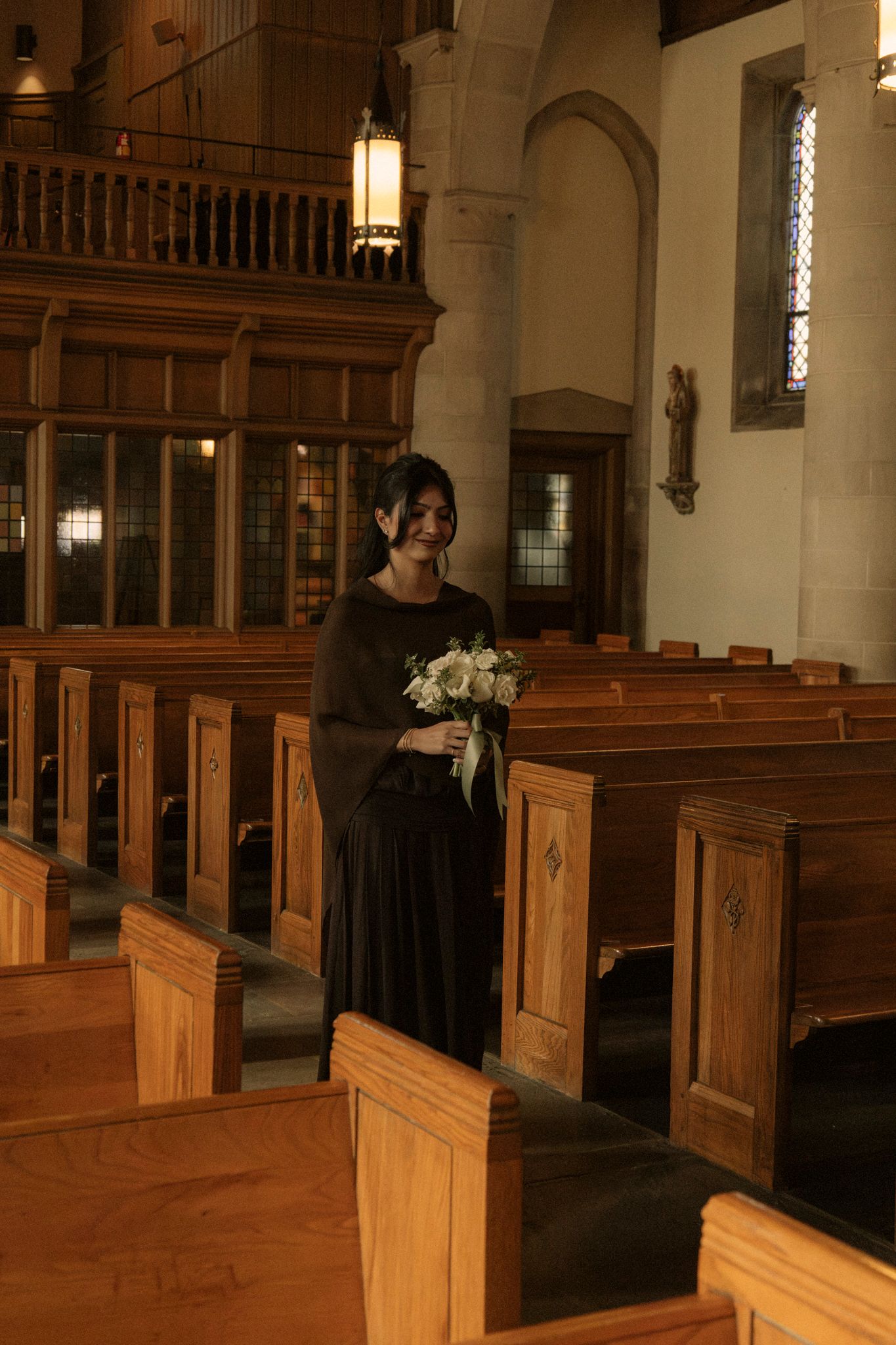 Smiling bridesmaid in black dress holds white floral bouquet while standing beside wooden church pews.