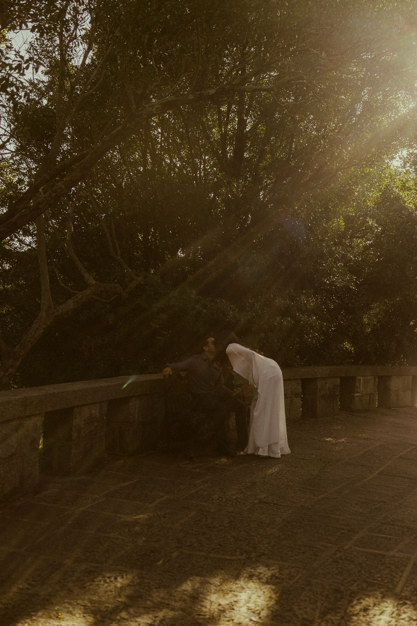 Bride in white dress leaning over stone bridge railing in golden sunlight surrounded by lush trees.