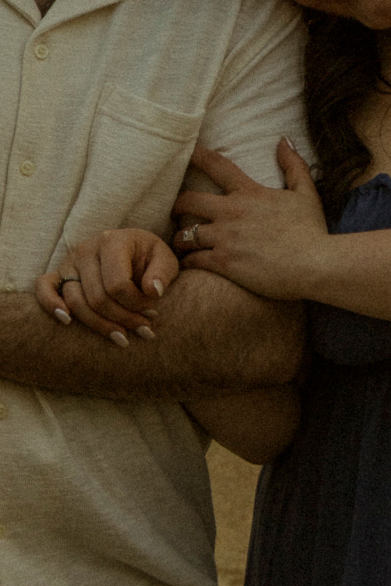 Close-up of a couple holding hands, woman wearing an engagement ring, dressed in cream and navy blue outfits.