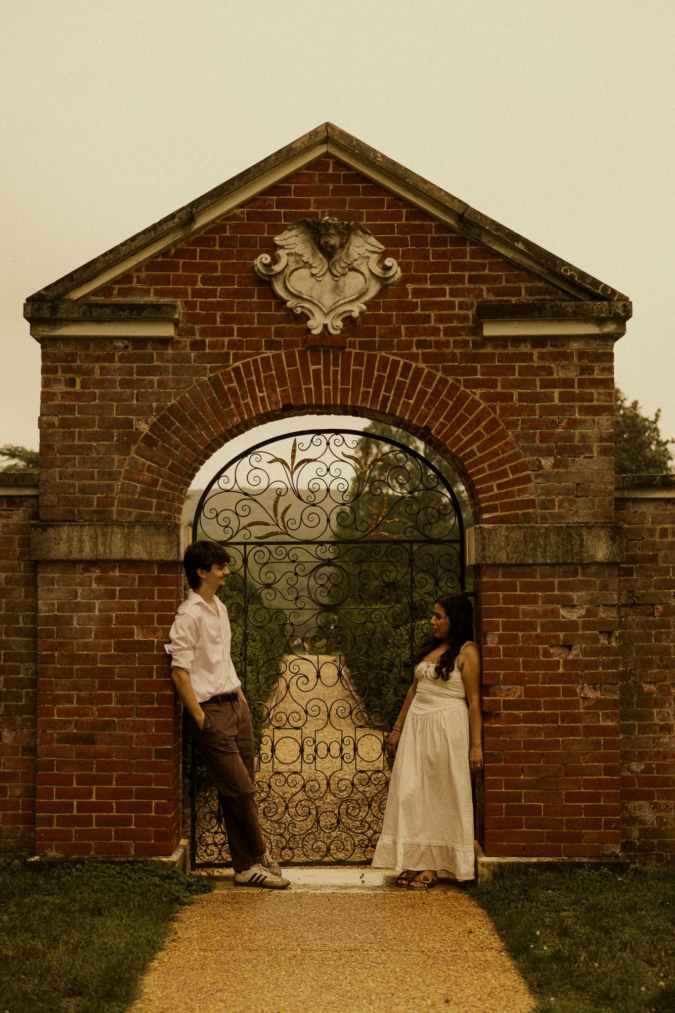 Couple standing in ornate brick archway with decorative iron gate, man in white shirt and woman in white dress facing each other.