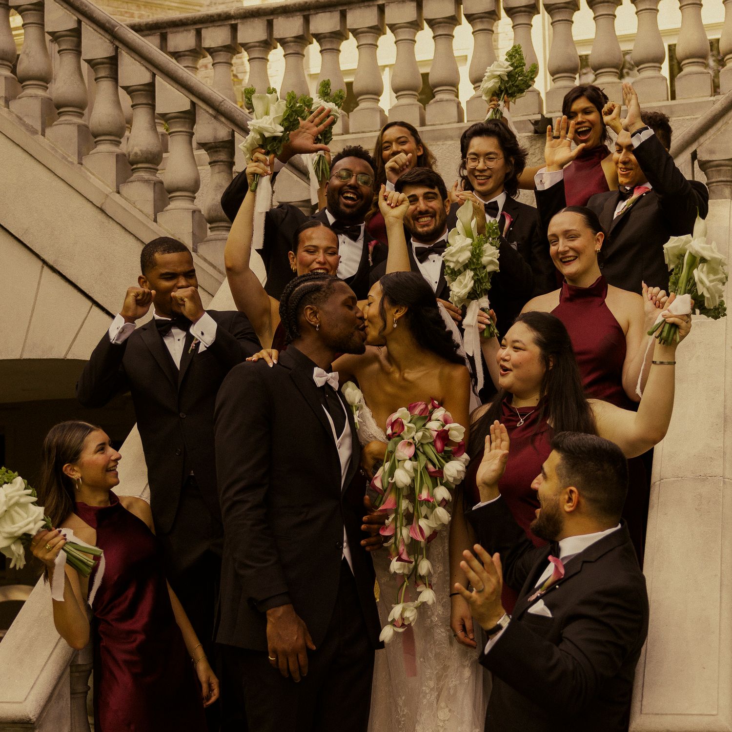 Wedding party celebrates on grand staircase as bride and groom share a kiss, bridesmaids in burgundy and groomsmen in black.