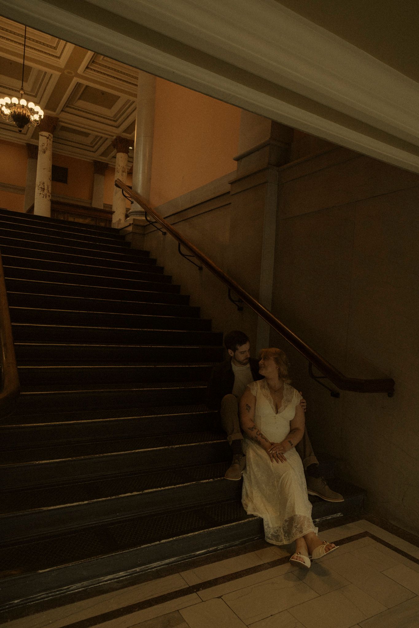 A figure in white sits on dark stairs in a dimly lit interior space with dramatic shadows and lighting.