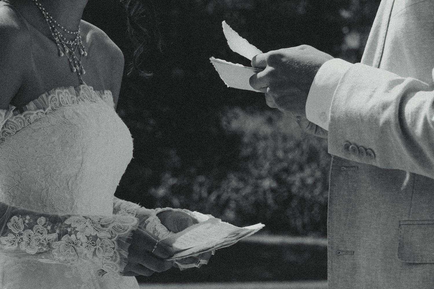 Black and white photo of a bride and groom exchanging wedding vows, holding papers during an outdoor ceremony.