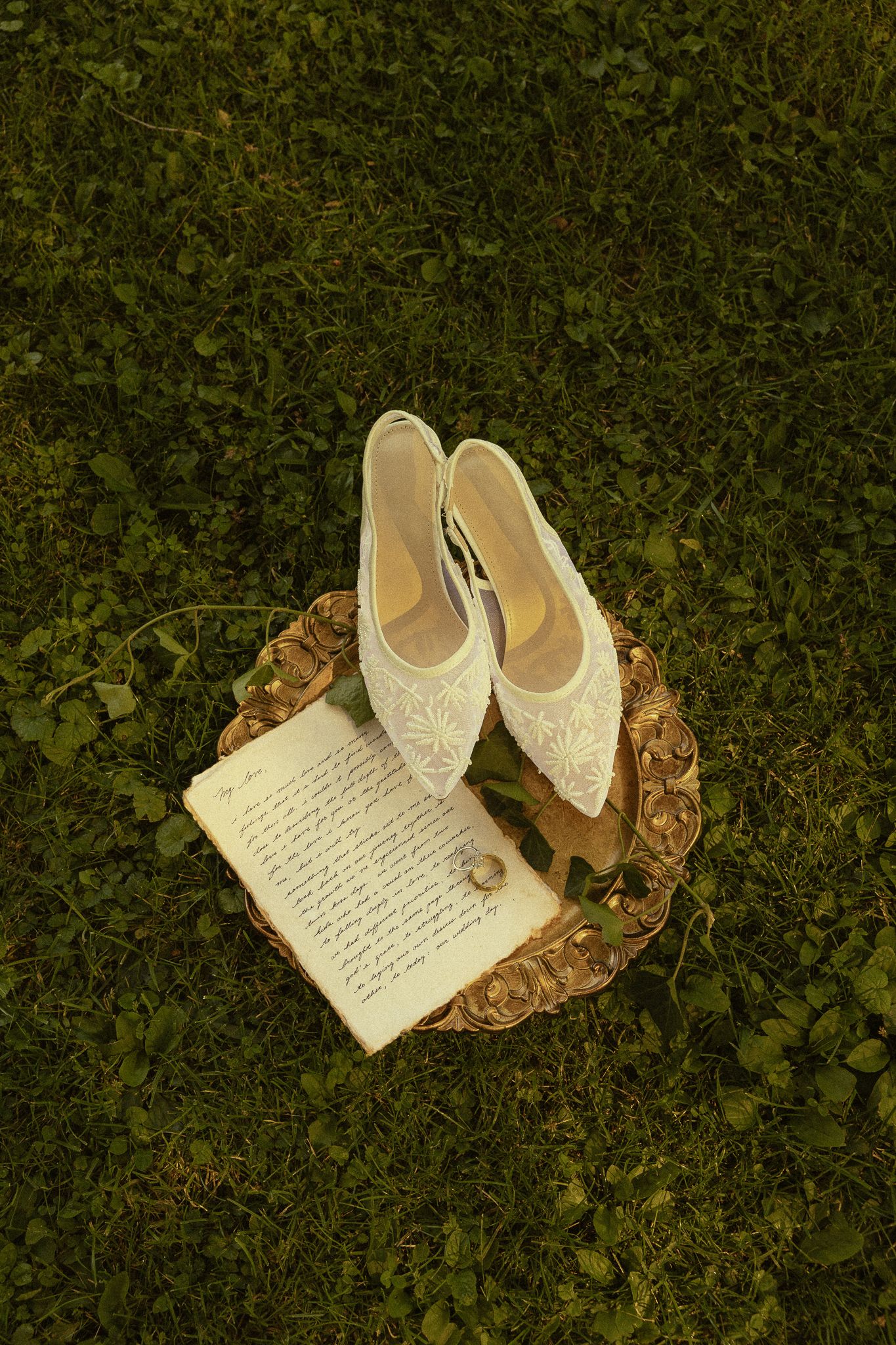 White lace bridal shoes and wedding rings on an open book atop a wicker tray, set on lush green grass.
