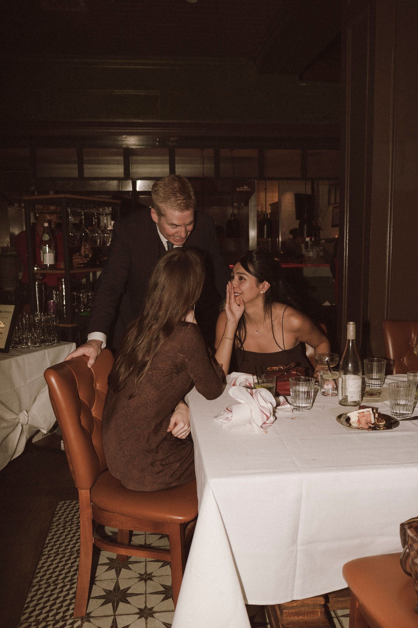 Man in suit leans over two women dining at elegant restaurant table with white tablecloth and wine.