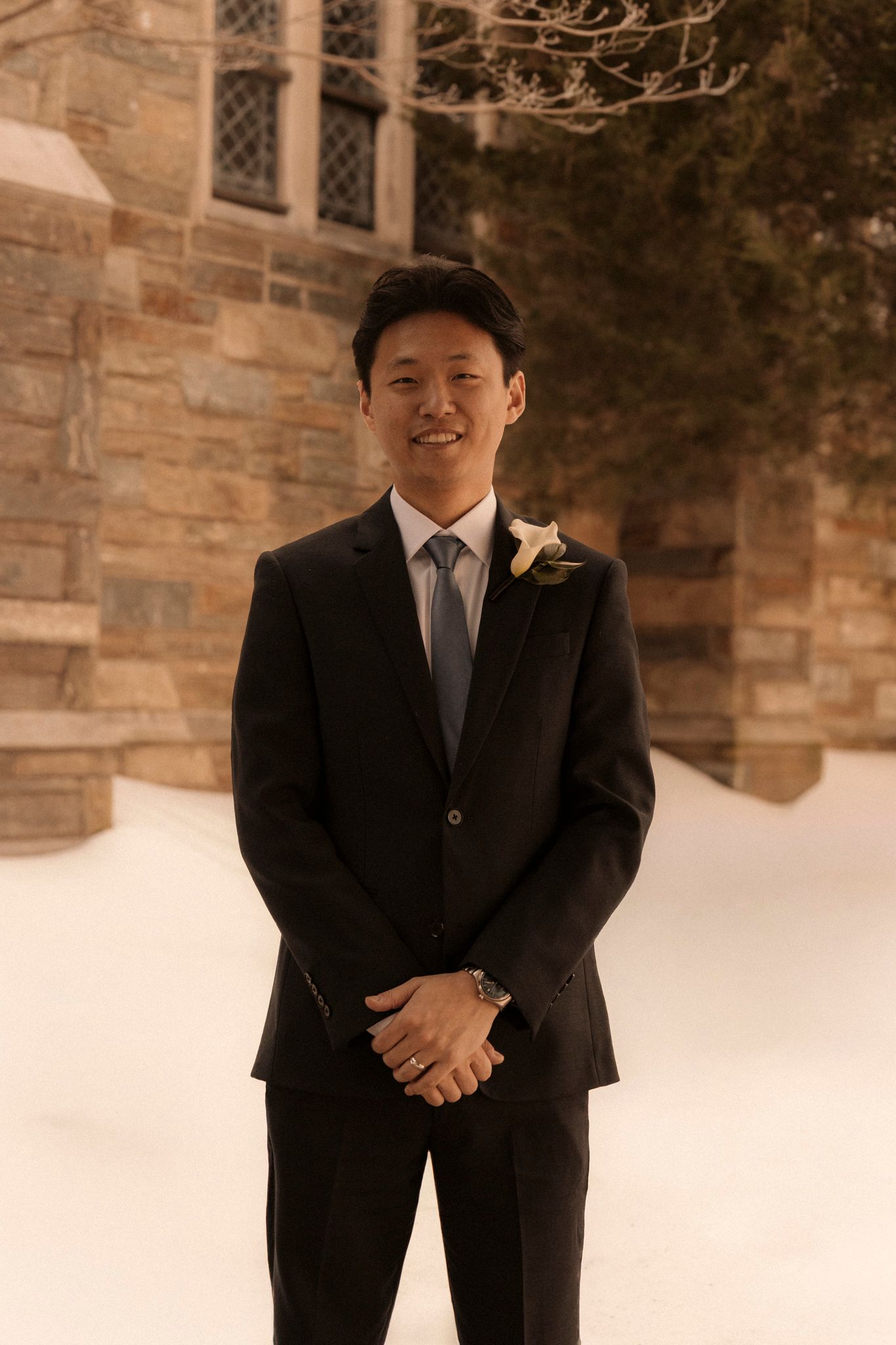 Smiling groom in black suit with boutonniere standing in snow outside a historic stone church.