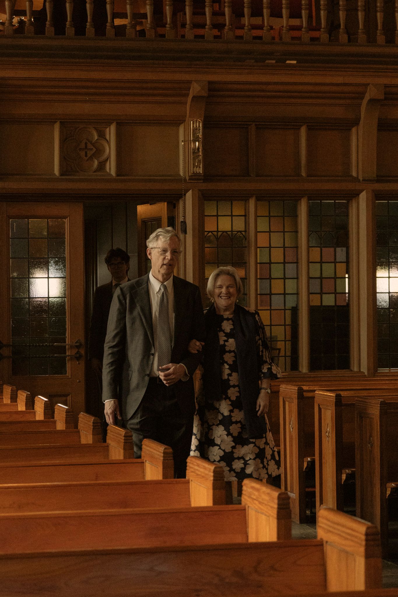 Elderly couple walking down church aisle past wooden pews with stained glass windows in background.