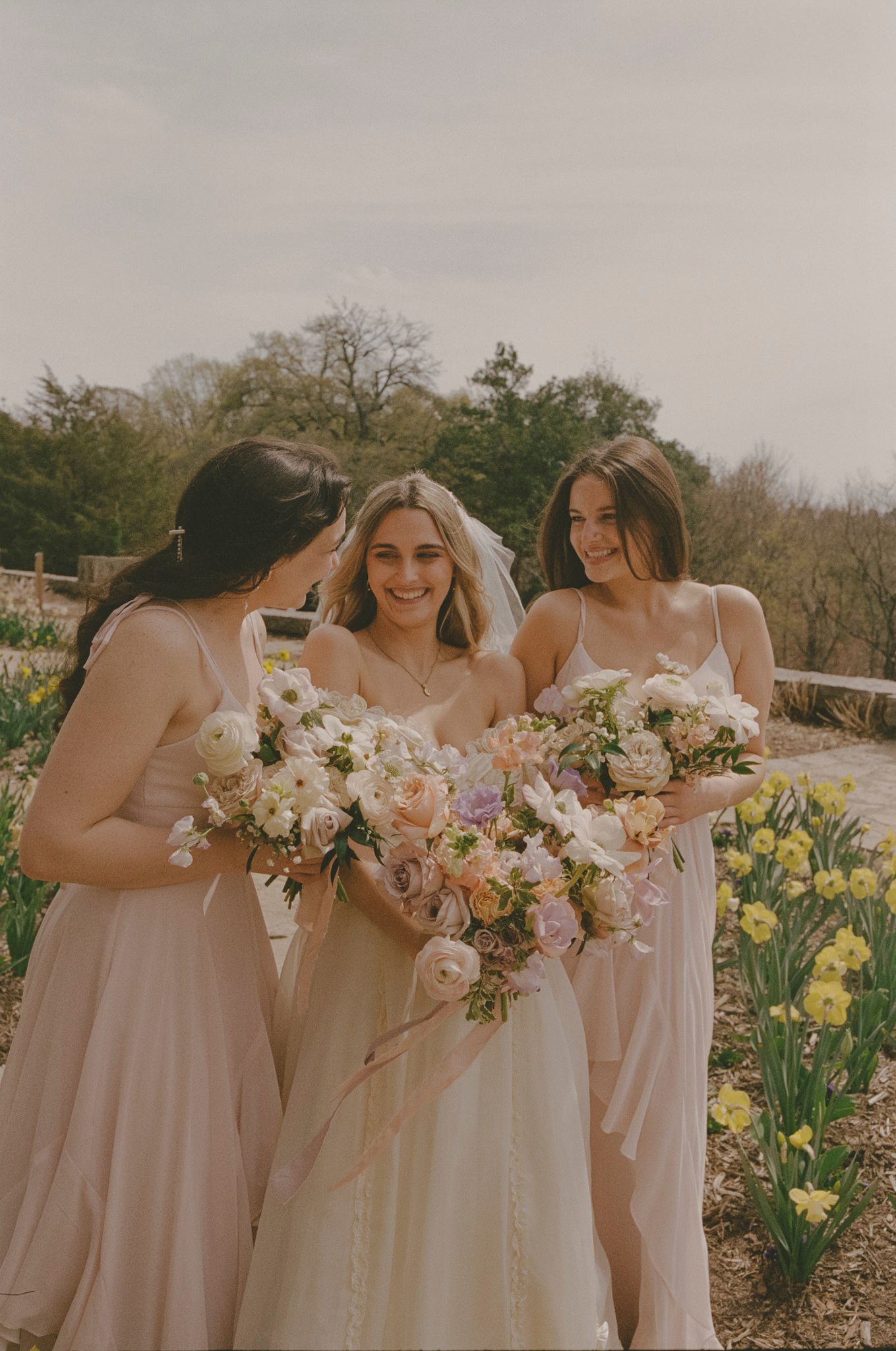 Bridesmaids in blush pink dresses pose together in a spring garden with blooming yellow daffodils.