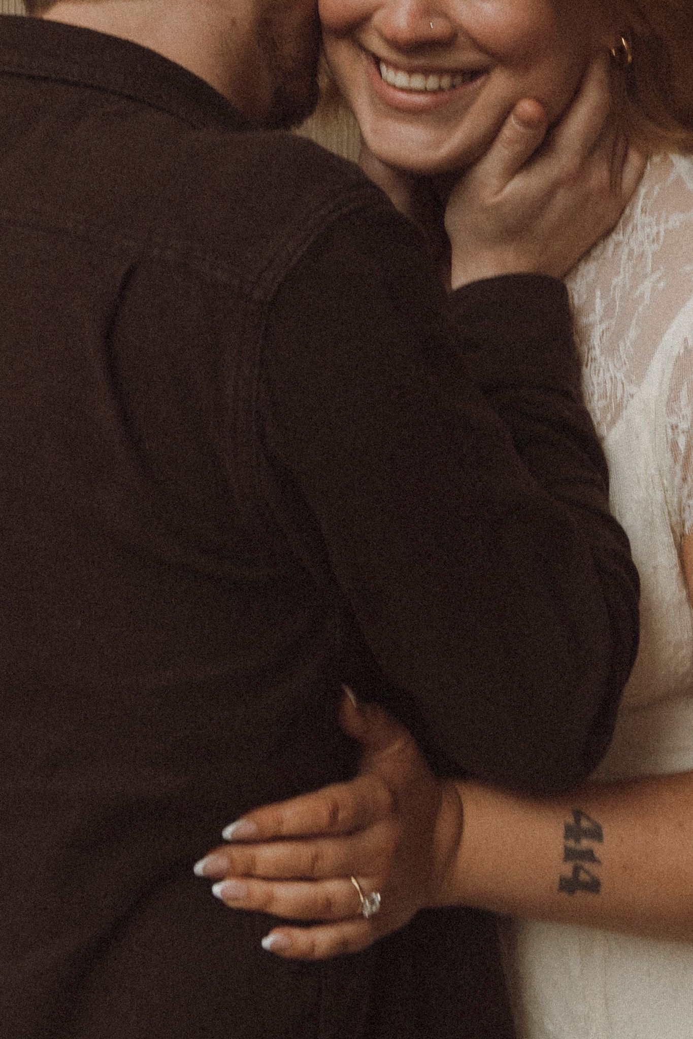 Close-up shot of a couple embracing, showing a white lace sleeve, small wrist tattoo and engagement ring.